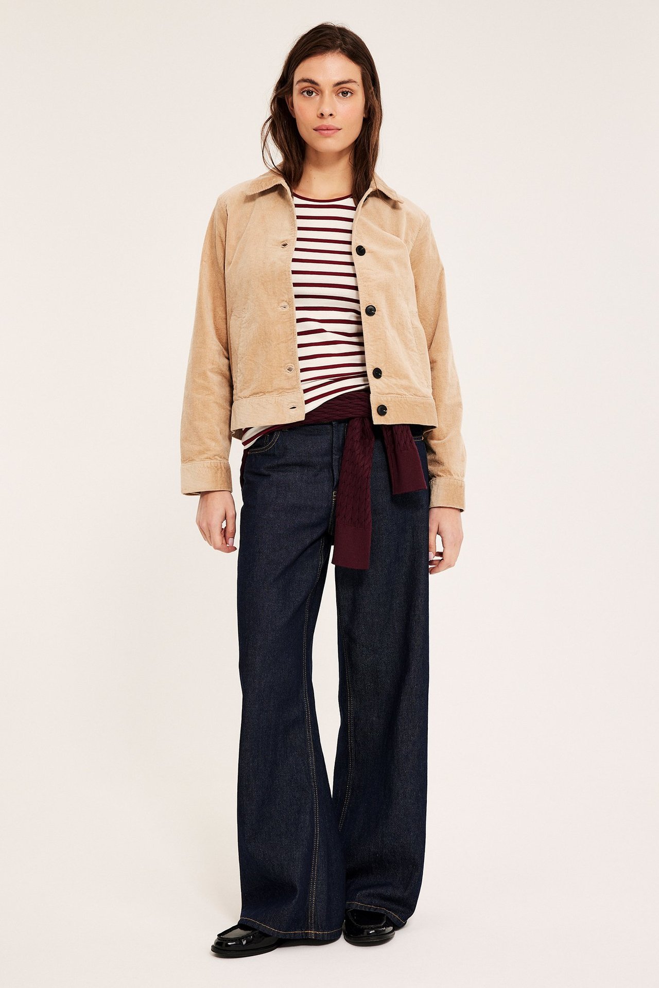 Frontbilde: Kvinne i hvit- og rødrandig kortermet topp, beige kordfløyelsjakke, mørkeblå jeans og svarte loafers.
