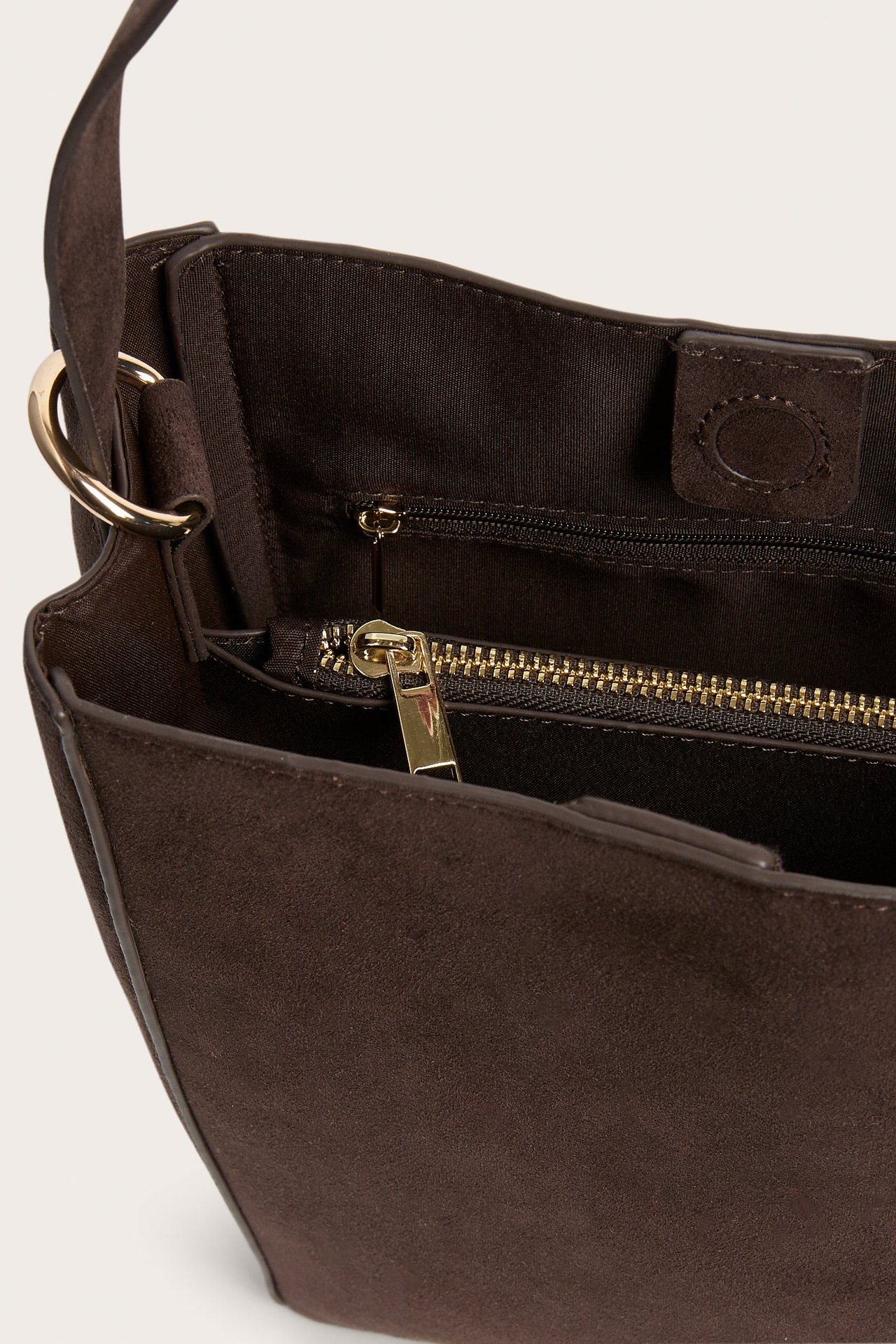 Nærbilde av en mørkebrun bøtteveske i fuskemocka med gullfarget innvendig glidelås og magnetlås.