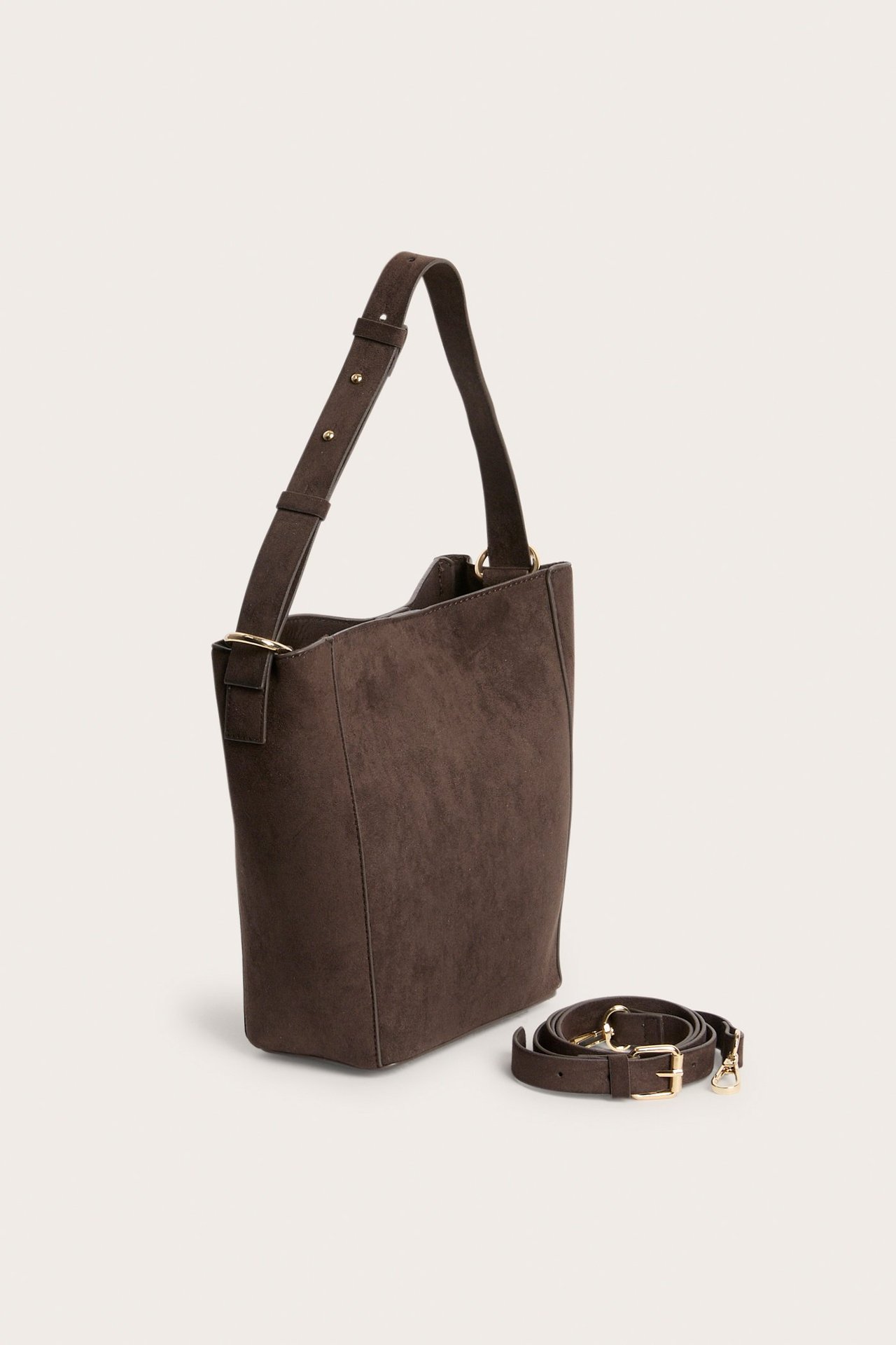 Vinklet visning av mørkebrun bøtteveske i fuskemocka med justerbar rem og separat skulderrem.