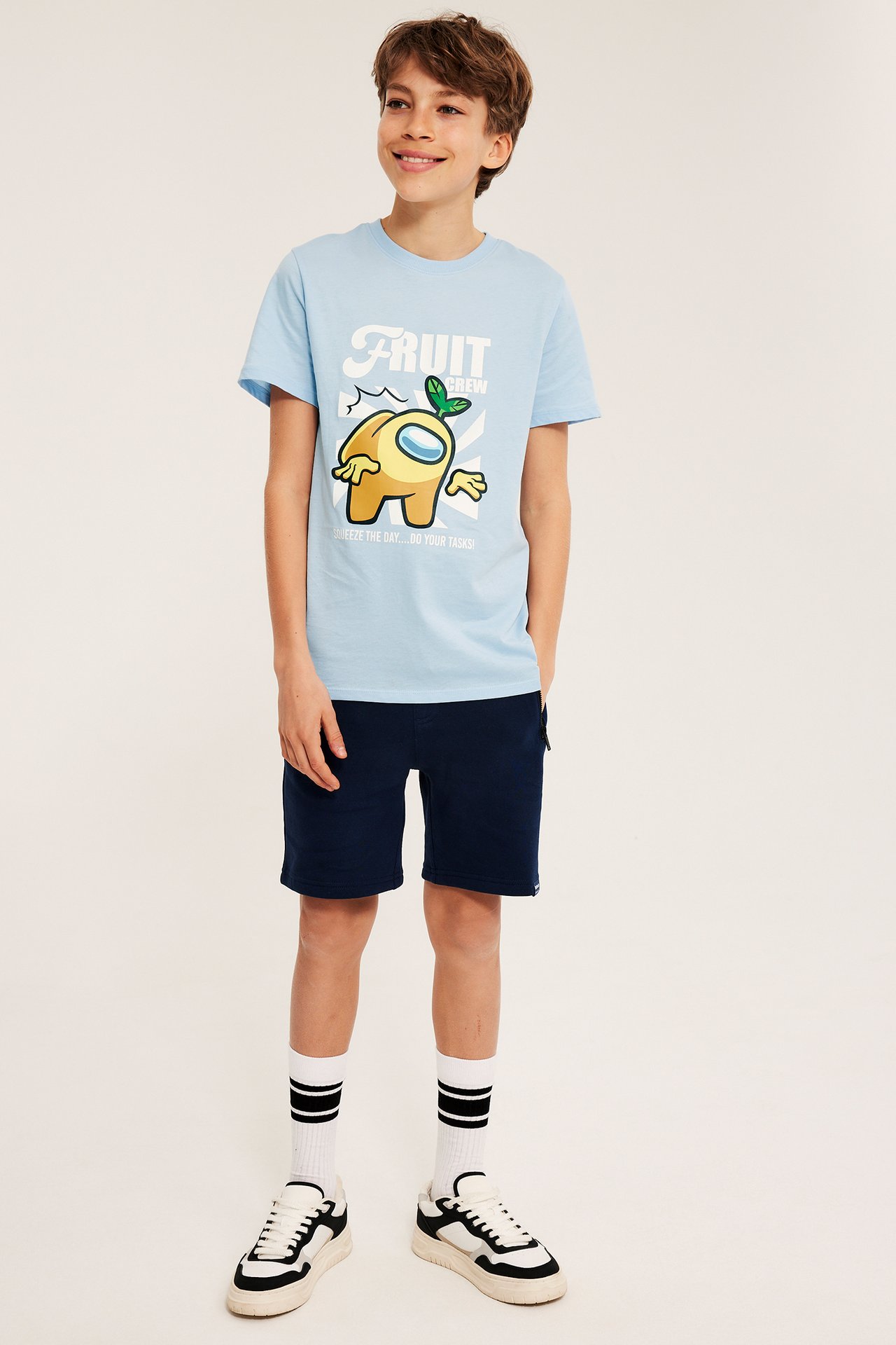 Edestä: Lapsi vaaleansinisessä Among Us -printti-T-paidassa, tummansinisissä shortseissa, valkoisissa raitasukissa ja lenkkareissa.