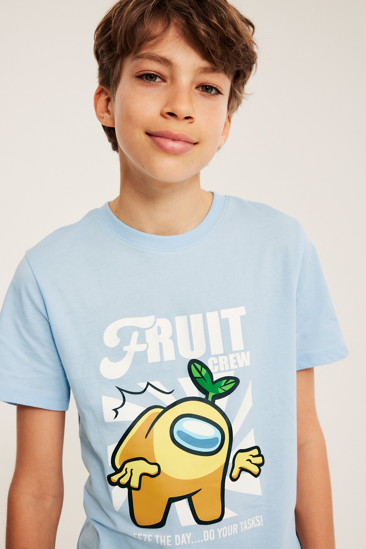 Widok z przodu: Dziecko w jasnoniebieskim bawełnianym t-shircie z żółtym nadrukiem Among Us 'Fruit Crew'.