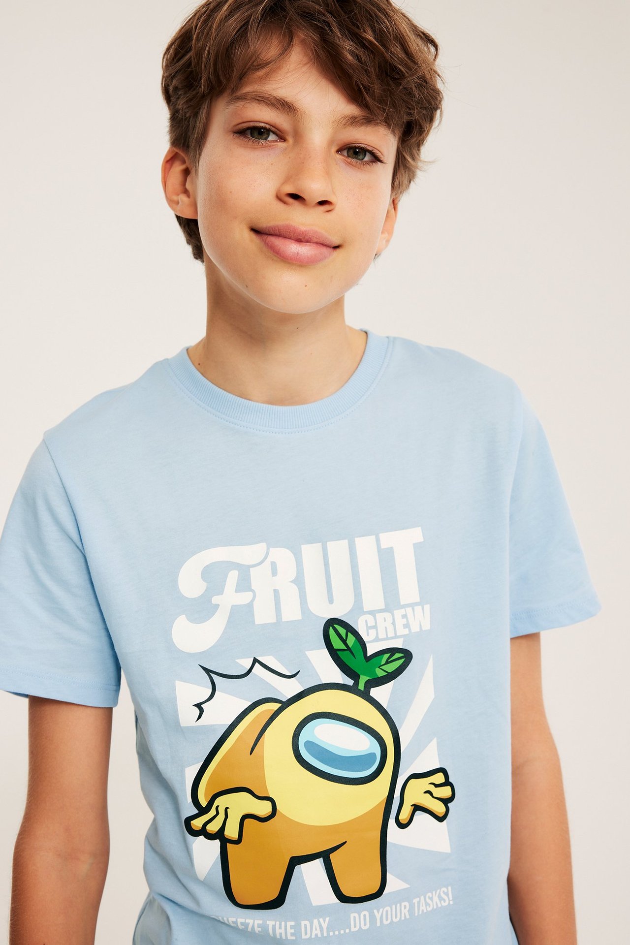 Widok z przodu: Dziecko w jasnoniebieskim bawełnianym t-shircie z żółtym nadrukiem Among Us 'Fruit Crew'.