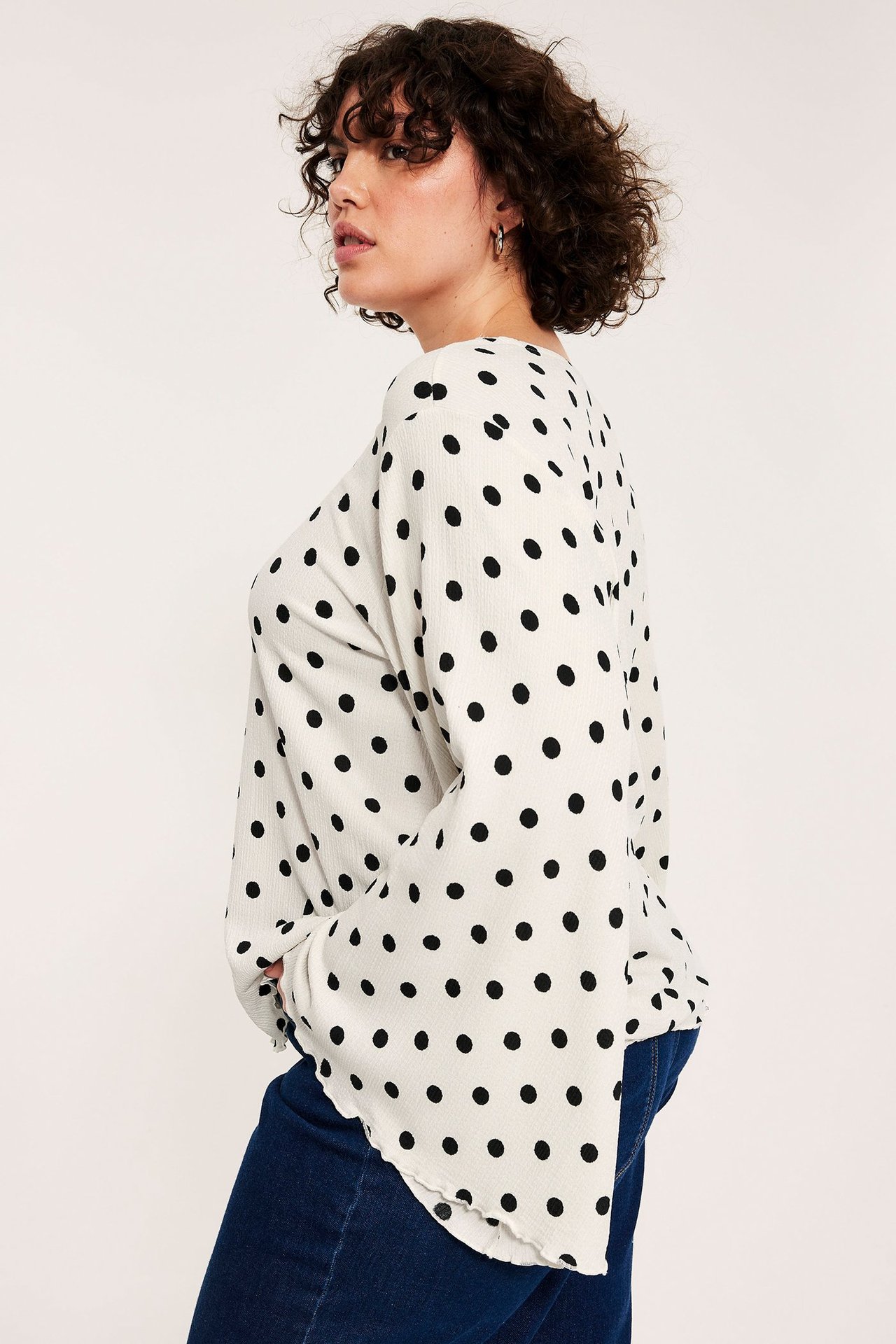 Widok z boku kobiety w bluzce off-white w kropki z szerokimi rękawami i granatowych dżinsach.