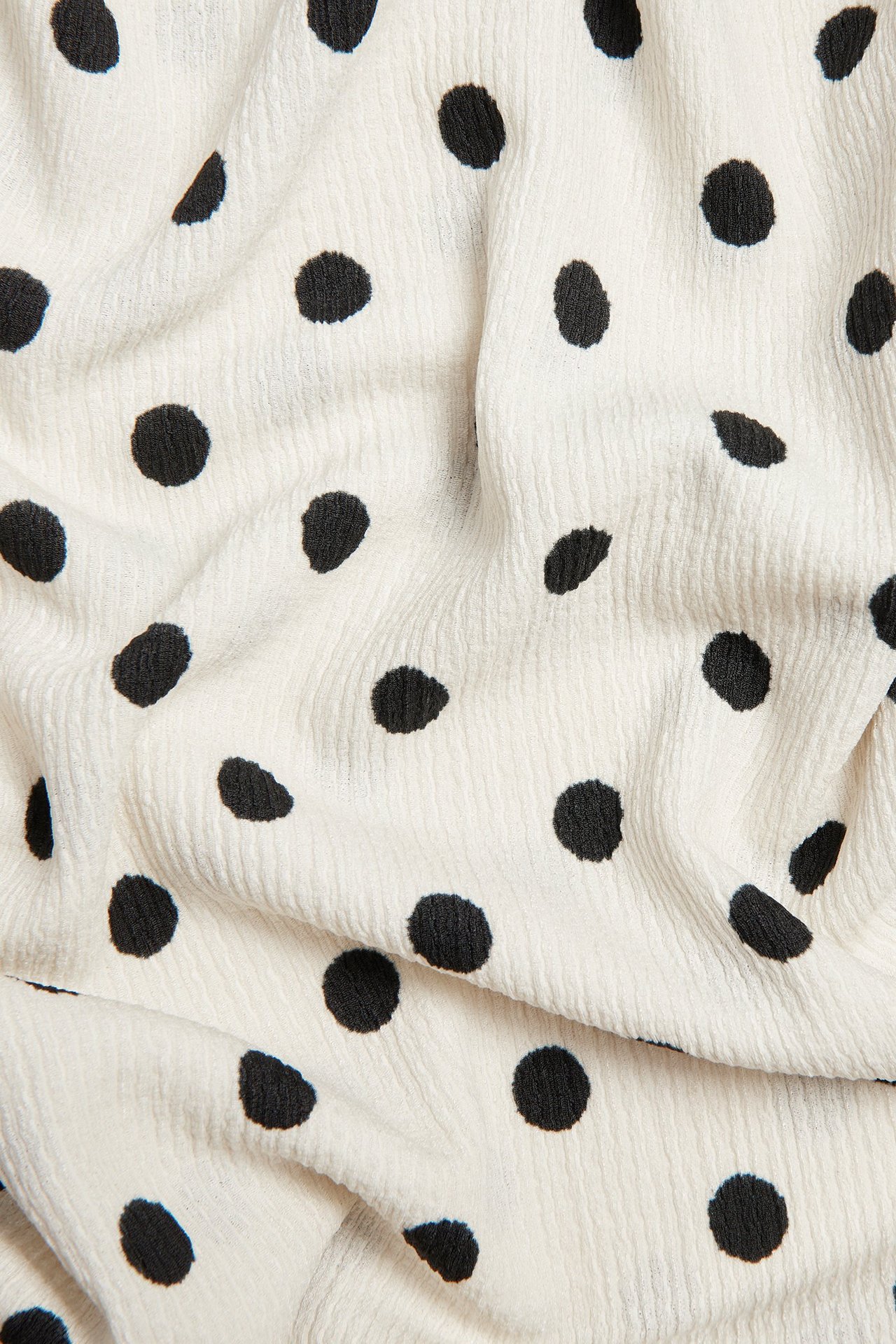 Nærbilde av offwhite teksturert blusestoff med svart prikkemønster og myke folder.