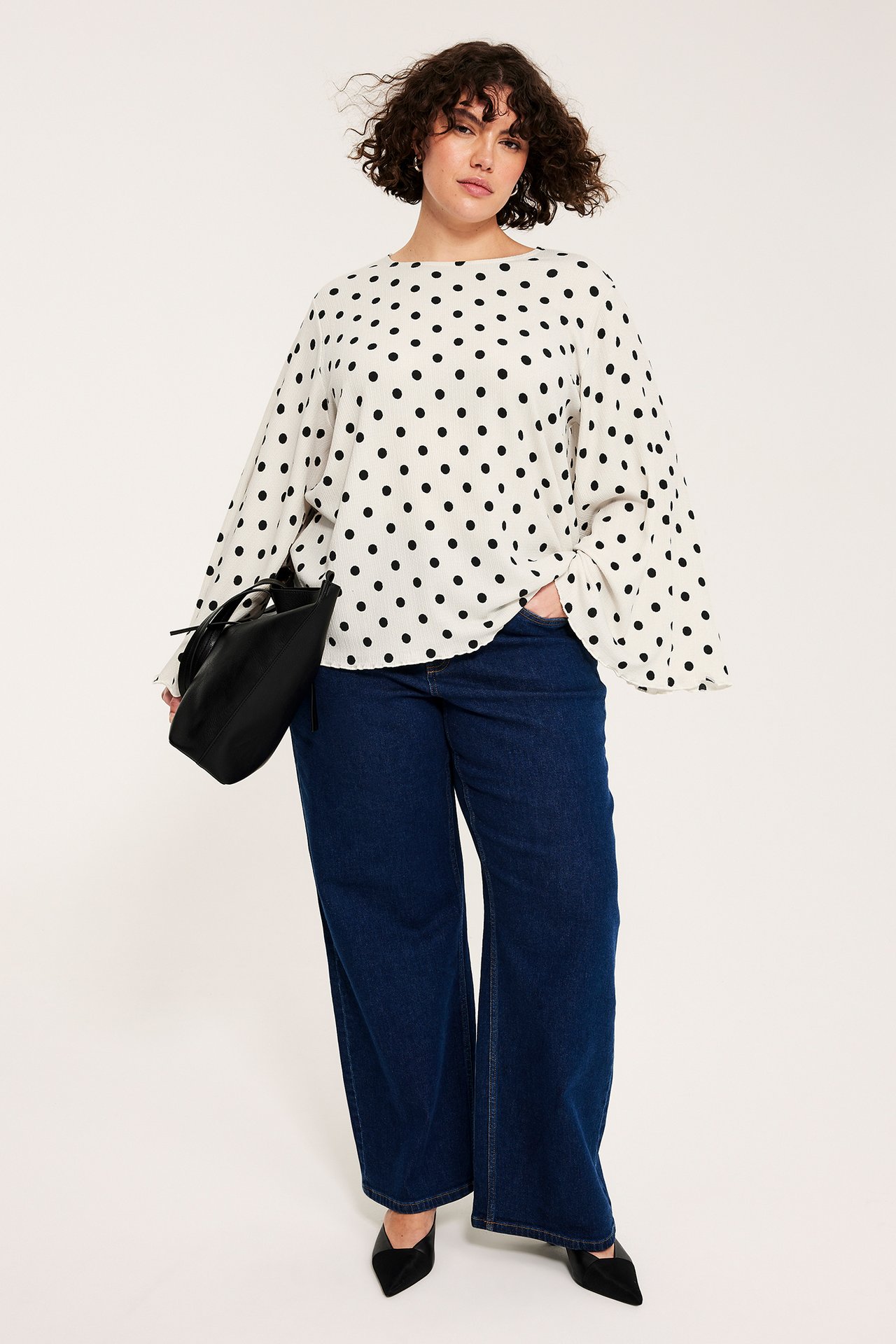Framifrån: Kvinna i offwhite prickig blus med vida ärmar, mörkblå vida jeans, svarta skor, hållande svart väska.
