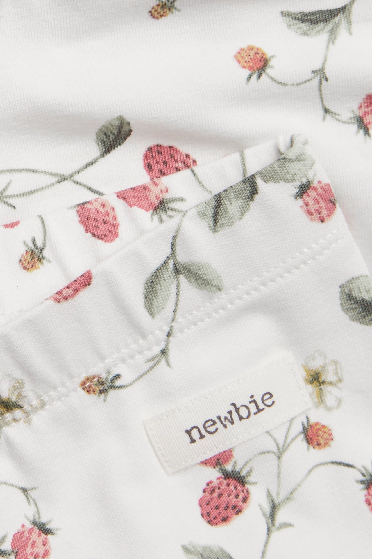 Zbliżenie na legginsy z dzianiny w kolorze ecru z czerwonym wzorem truskawkowym i widoczną metką 'newbie'.