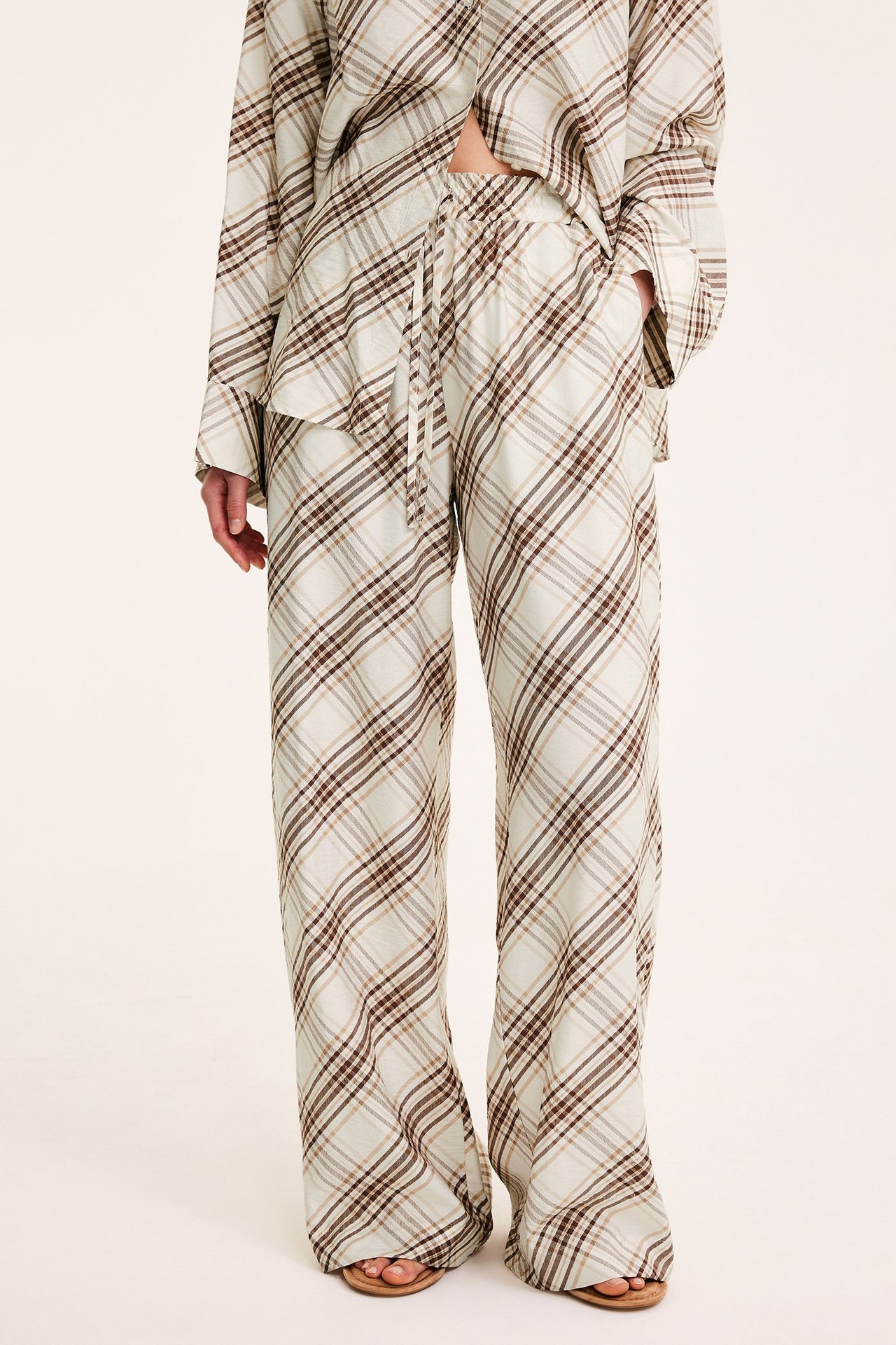 Naisen edestä: leveät beigeruutuiset housut kiristysnyörillä vyötäröllä, yhteensopiva paita ja ruskeat sandaalit.