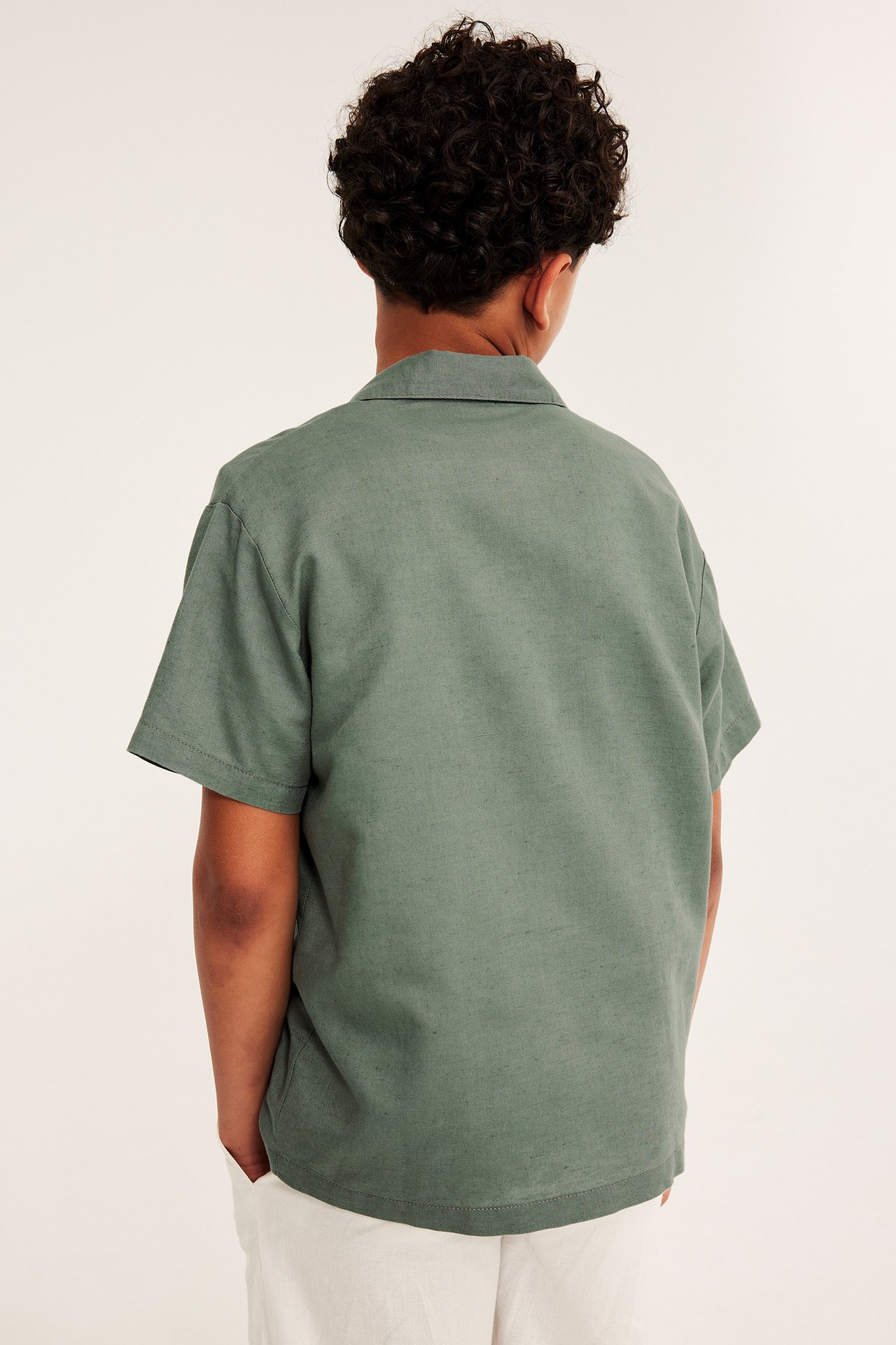 Lapsi selin vihreässä lyhythihaisessa pellavasekoitepaidassa ja vaaleissa housuissa.