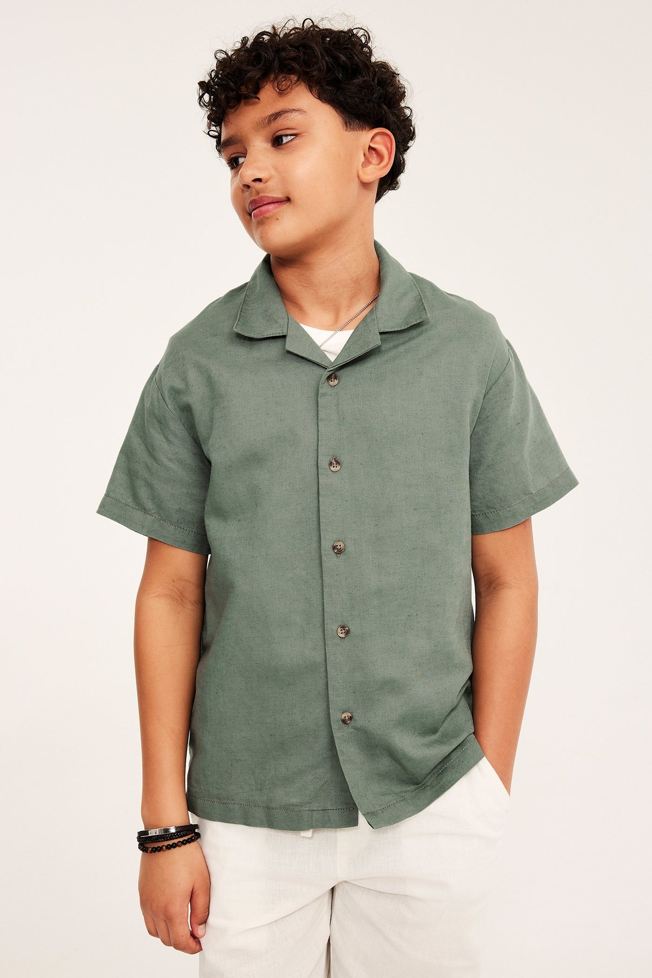 Framifrån, en pojke i grön kortärmad skjorta i linneblandning, vita shorts och svart armband.