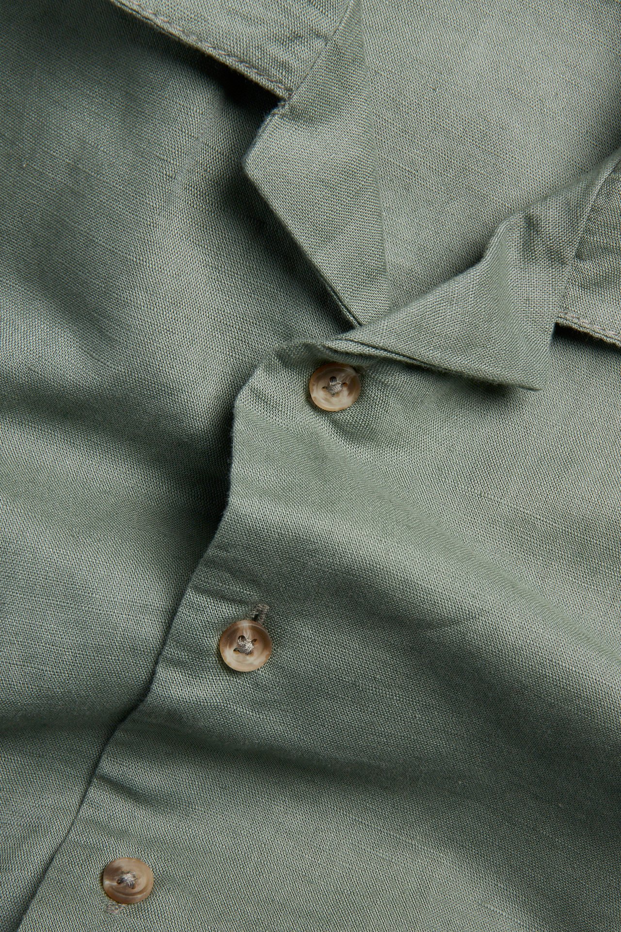 Närbild av en grön kortärmad skjorta i linneblandning med krage och bruna knappar.