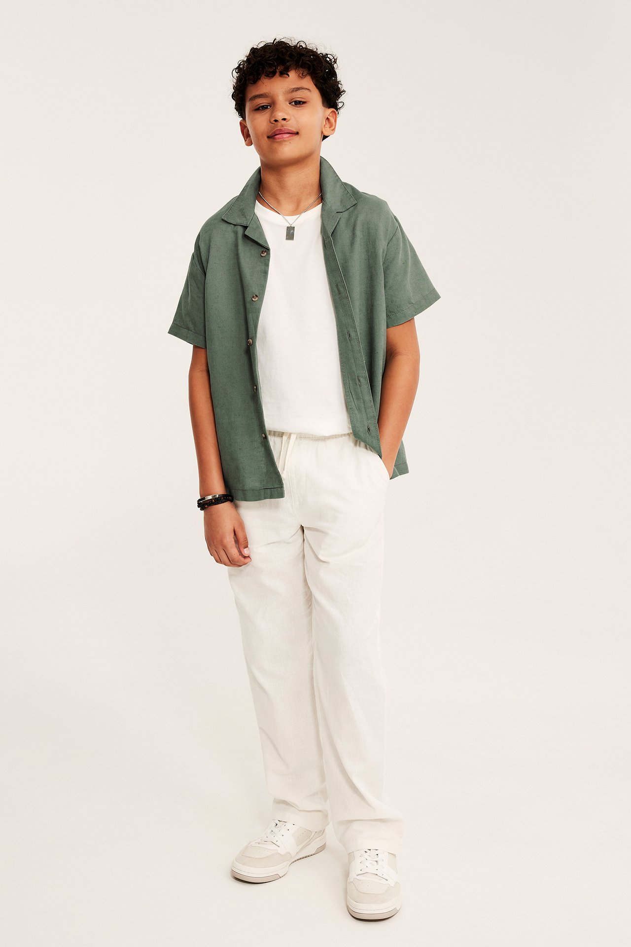 Etunäkymä: Lapsi vihreässä pellavasekoitepaidassa, valkoinen t-paita, valkoiset housut ja lenkkarit.