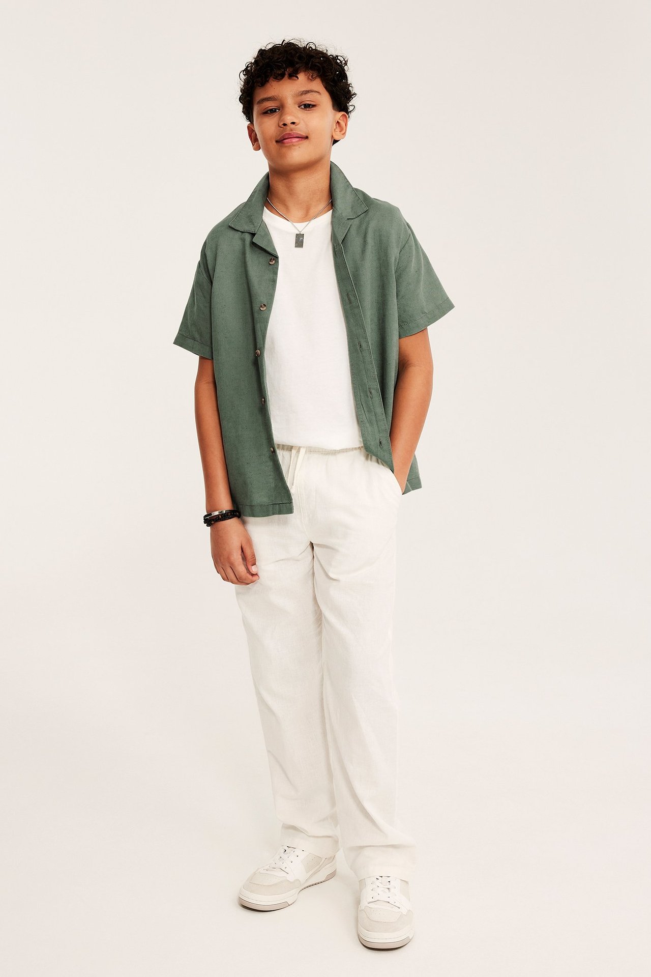 Widok z przodu: Dziecko w zielonej koszuli lnianej z krótkim rękawem, białym T-shircie, spodniach i trampkach.