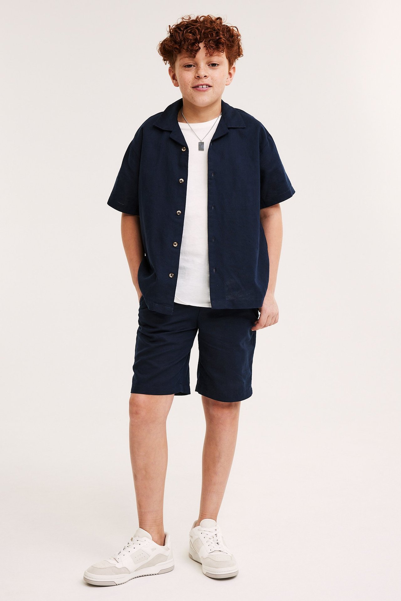 Helbild framifrån: Barn i mörkblå kortärmad linneskjorta över vit t-shirt, mörkblå shorts, vita sneakers.