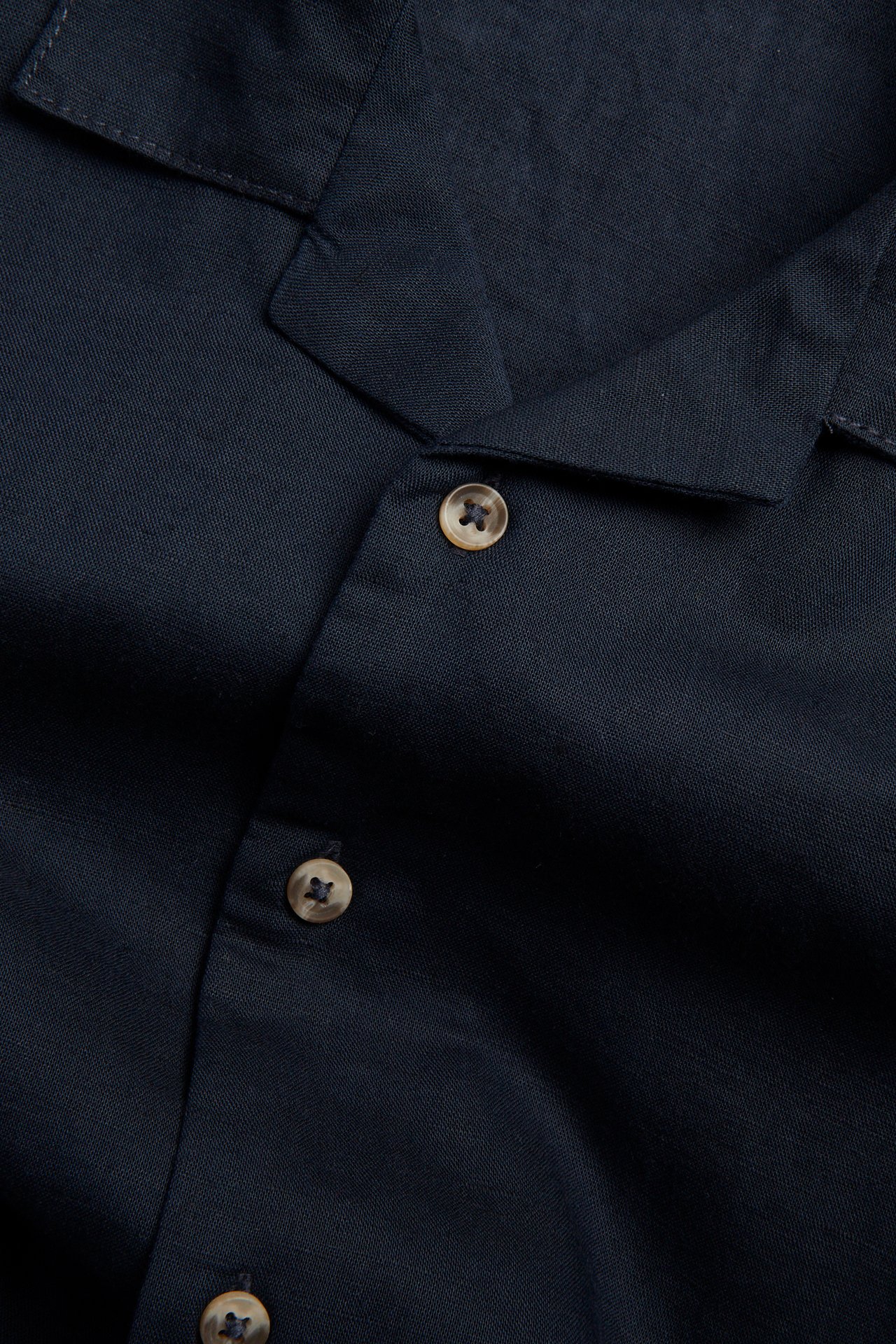 Närbild av en mörkblå kortärmad skjorta i linneblandning med ljusbruna knappar.