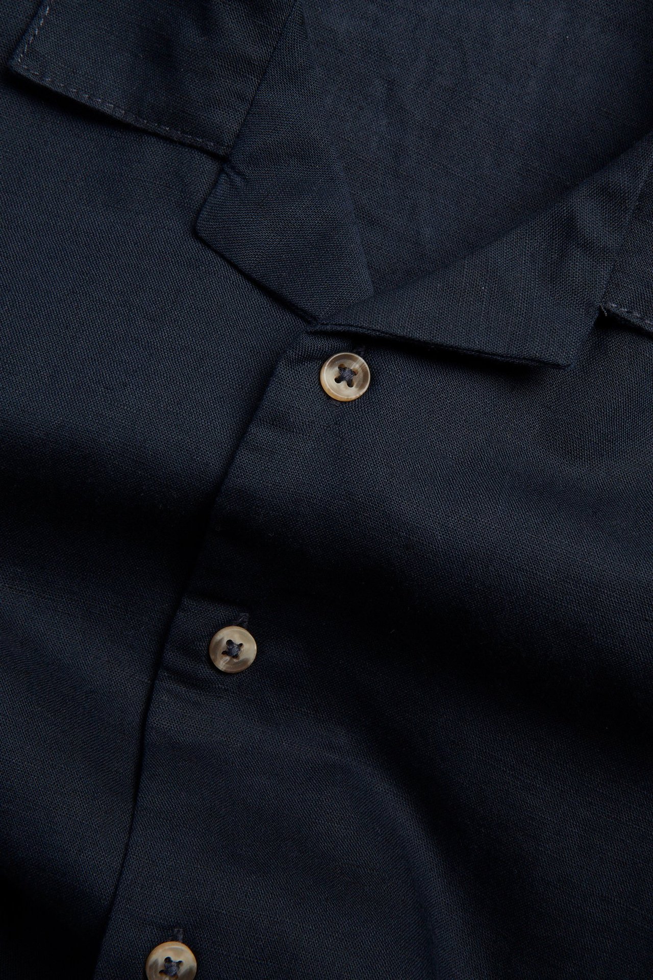 Närbild av en mörkblå kortärmad skjorta i linneblandning med ljusbruna knappar.