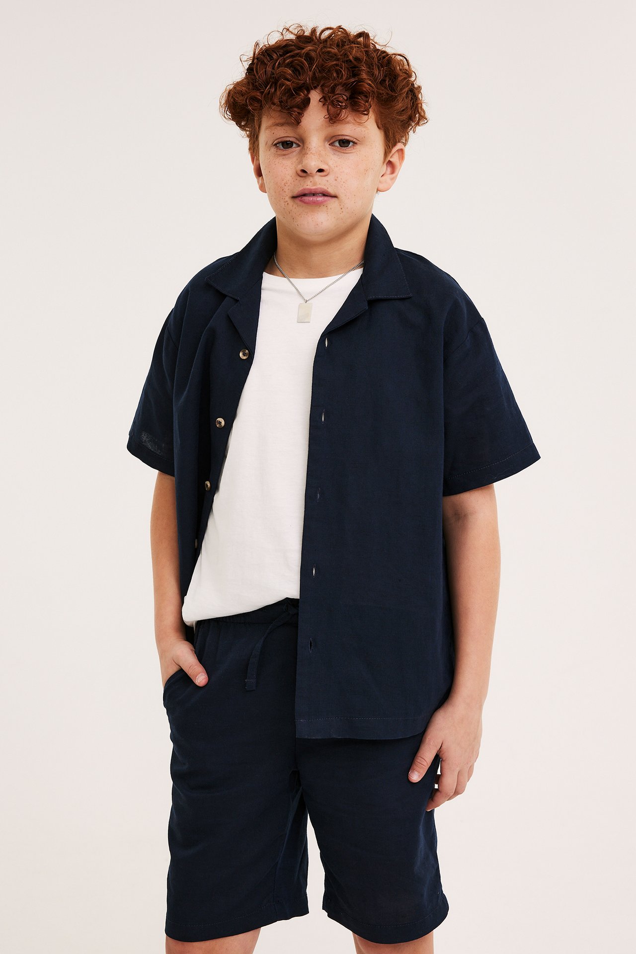 Barn, framifrån: Mörkblå kortärmad linneblandningsskjorta, matchande shorts, vit t-shirt och silverhalsband.