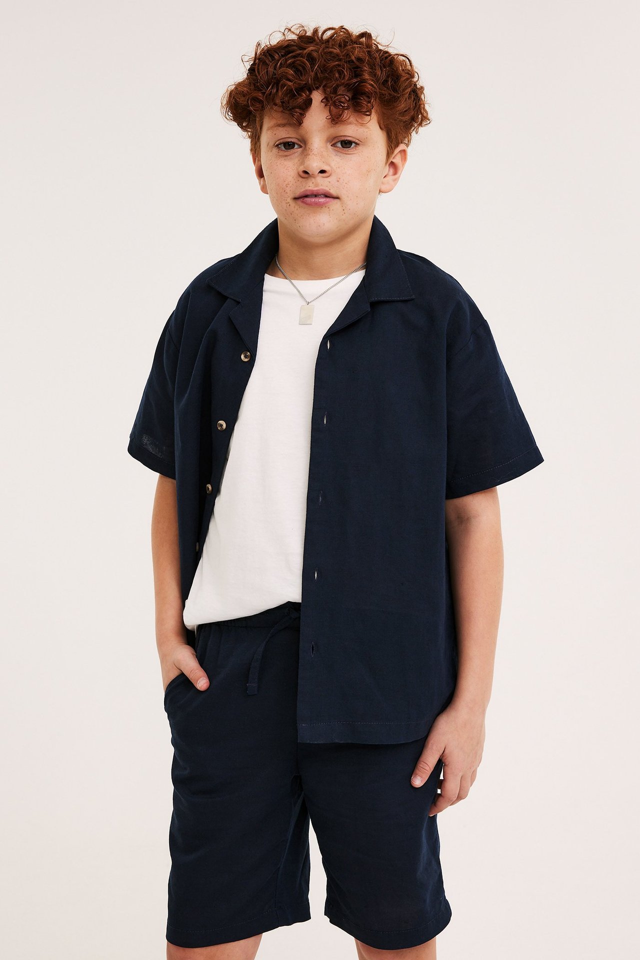 Barn, framifrån: Mörkblå kortärmad linneblandningsskjorta, matchande shorts, vit t-shirt och silverhalsband.