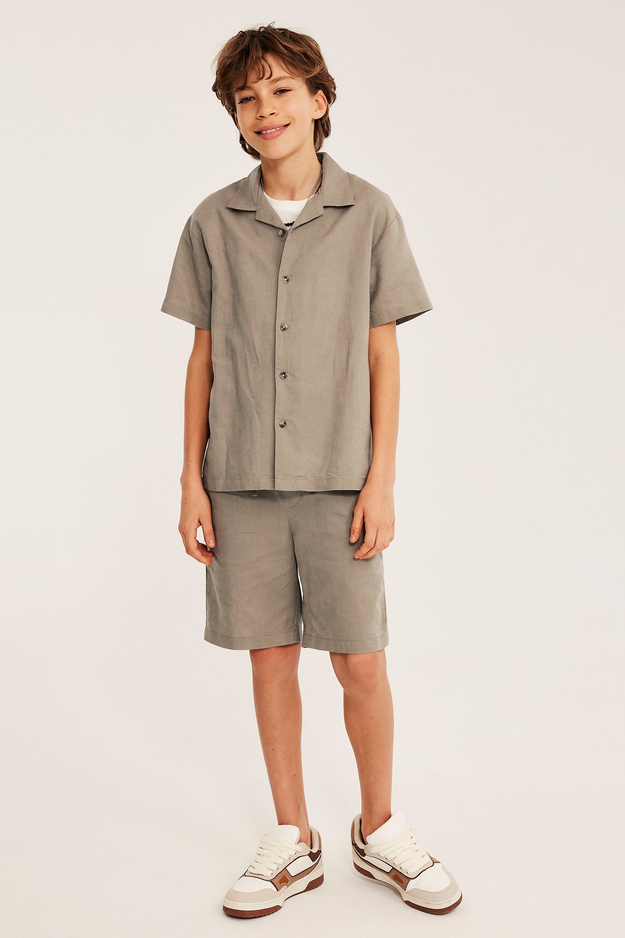 Framifrån: Leende barn i beige kortärmad linneskjorta, över vit t-shirt och matchande shorts.