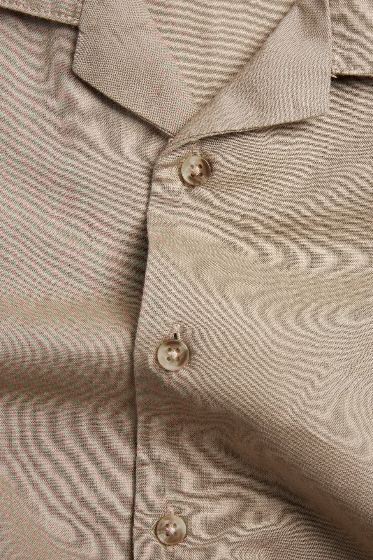 Närbild av en beige kortärmad skjorta i linneblandning, som visar knappslå med knappar och krage.