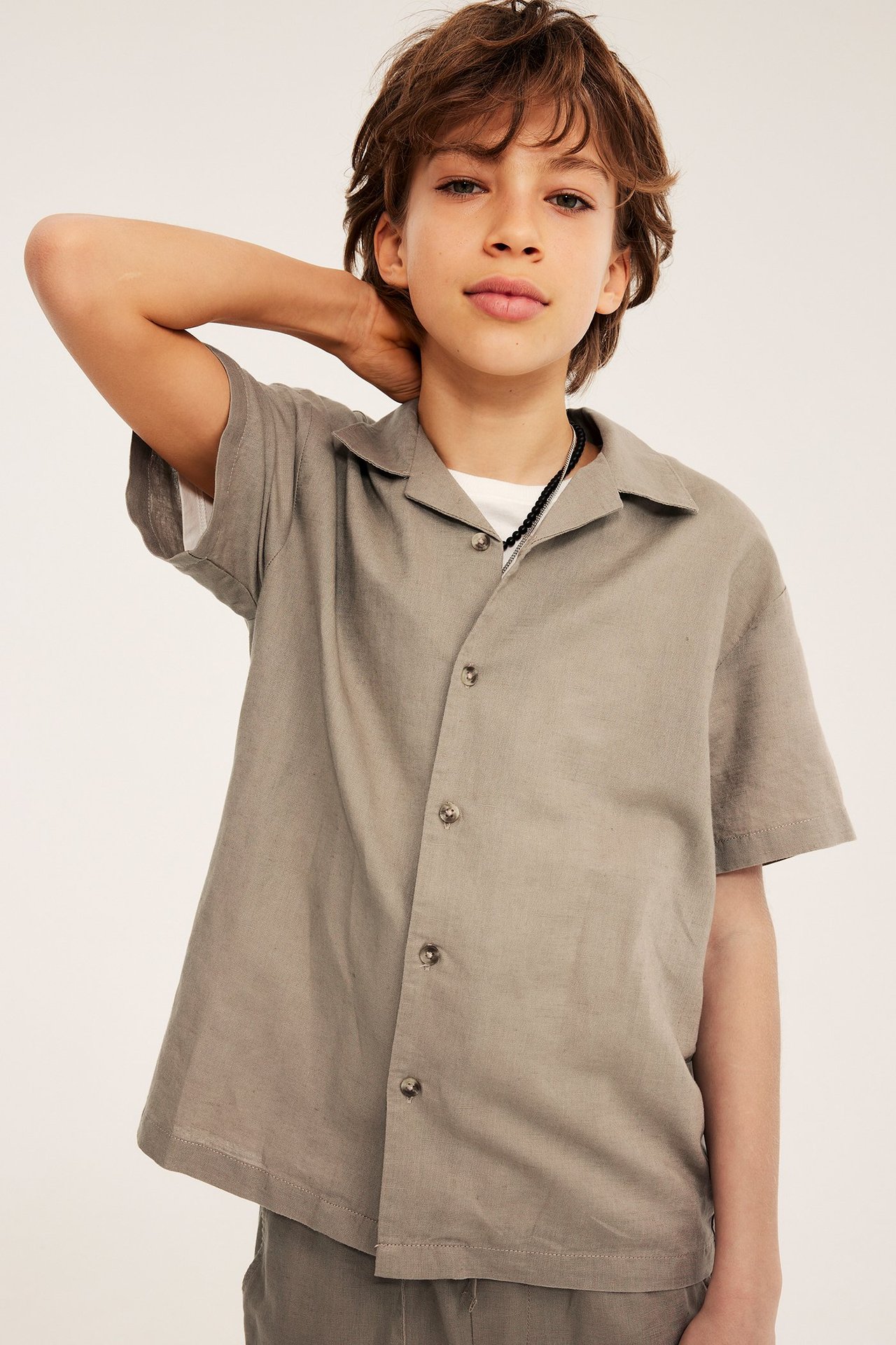 Framsida: Barn i beige kortärmad linneskjorta, vit t-shirt, mörka shorts och svart halsband.