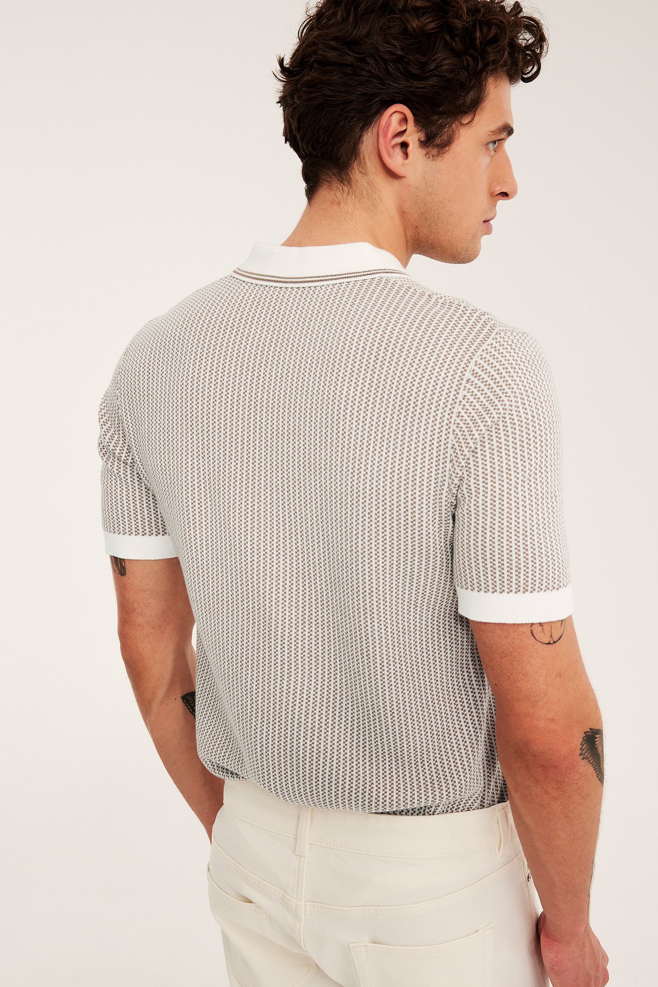 Man i sidovy bär beige jacquardmönstrad stickad pikétröja med vit krage/muddar och ljusbeige byxor.