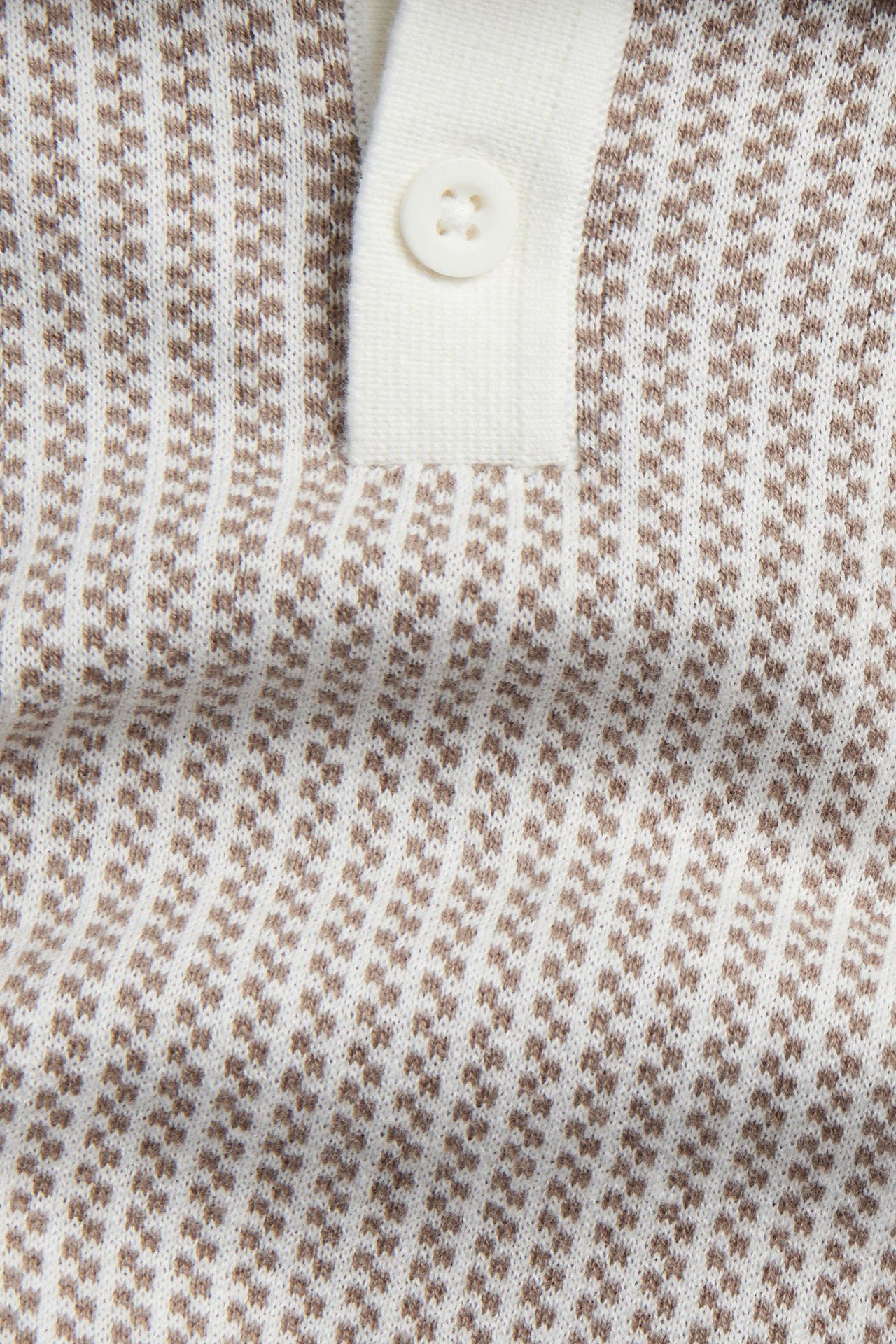Lähikuva beigestä ja luonnonvalkoisesta kuviolliseksi neulotusta pikeepaidasta, jossa näkyy nappi nappilistassa.