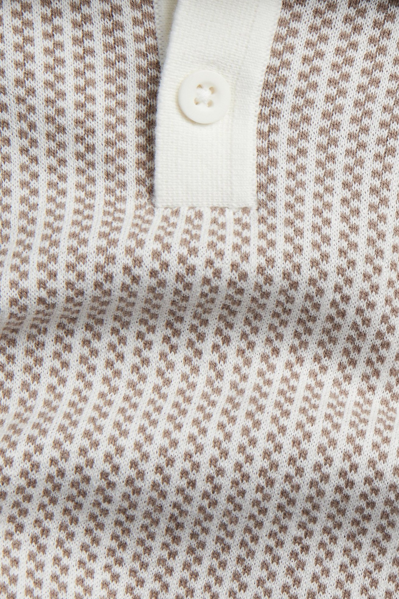 Nærbilde av en beige og naturhvit mønsterstrikket pikéskjorte med én synlig knapp på knappelukningen.