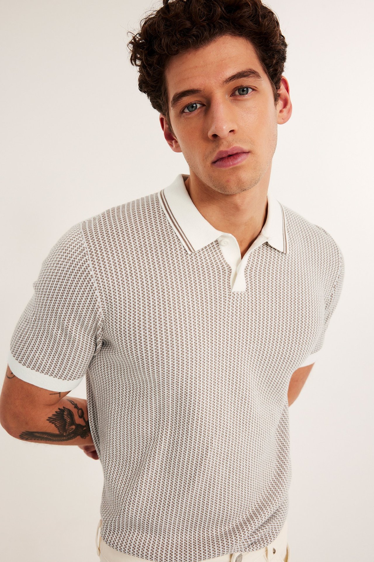 Framifrån på man i beige jacquardstickad pikétröja med vit krage, bruna ränder, två knappar.