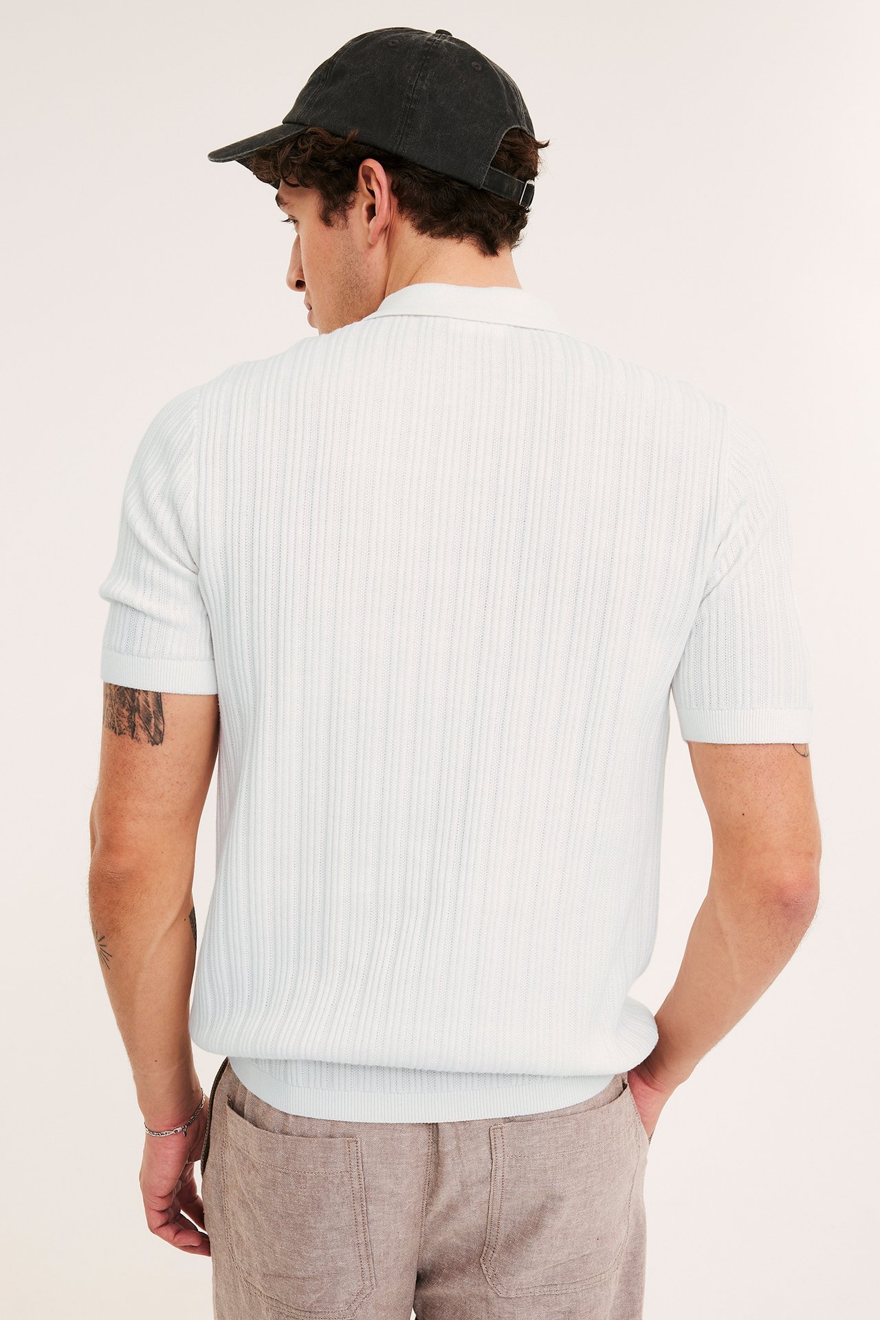 Mężczyzna tyłem w kremowej prążkowanej koszulce polo z krótkim rękawem, ciemnoszarej czapce i brązowych spodniach.