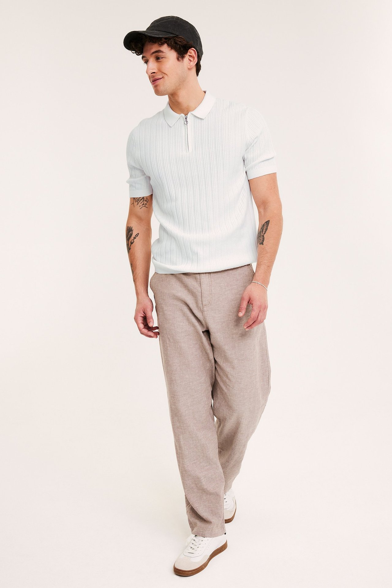 Edestä: mies yllään luonnonvalkoinen ribbineulottu half-zip pikeepaita, beiget housut, valkoiset tennarit, musta lippalakki.