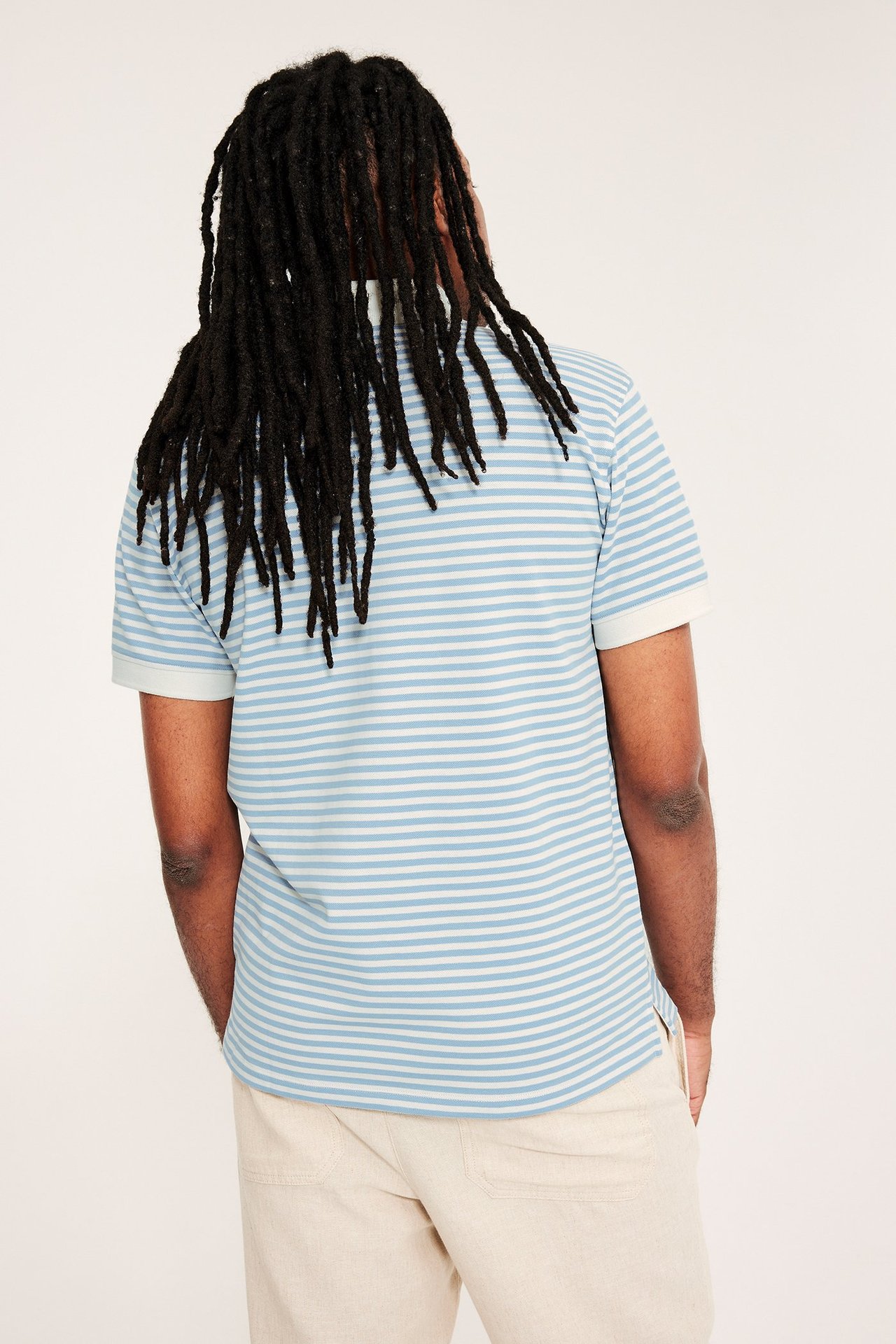 Takakuva miehestä, yllään vaaleansinivalkoinen raidallinen puuvillapikeepaita ja vaaleanbeiget housut.