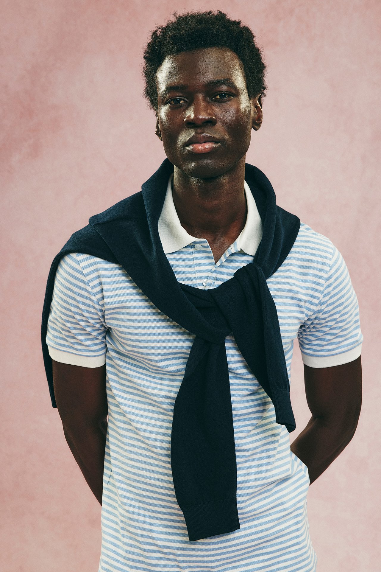 Widok z przodu: Mężczyzna w jasnoniebieskim, pasiastym bawełnianym polo i granatowym swetrze na ramionach.