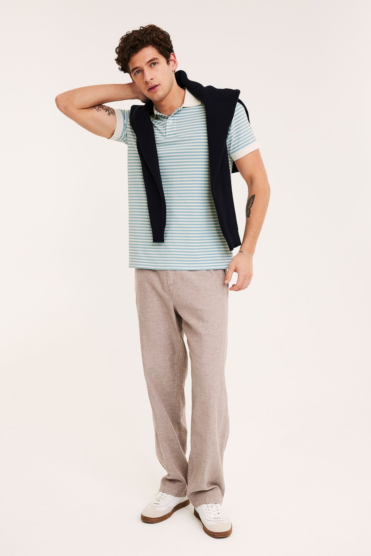 Front view: Man in light blue randig bomullspiké, marinblå tröja på axlarna, beiga byxor, vita sneakers.