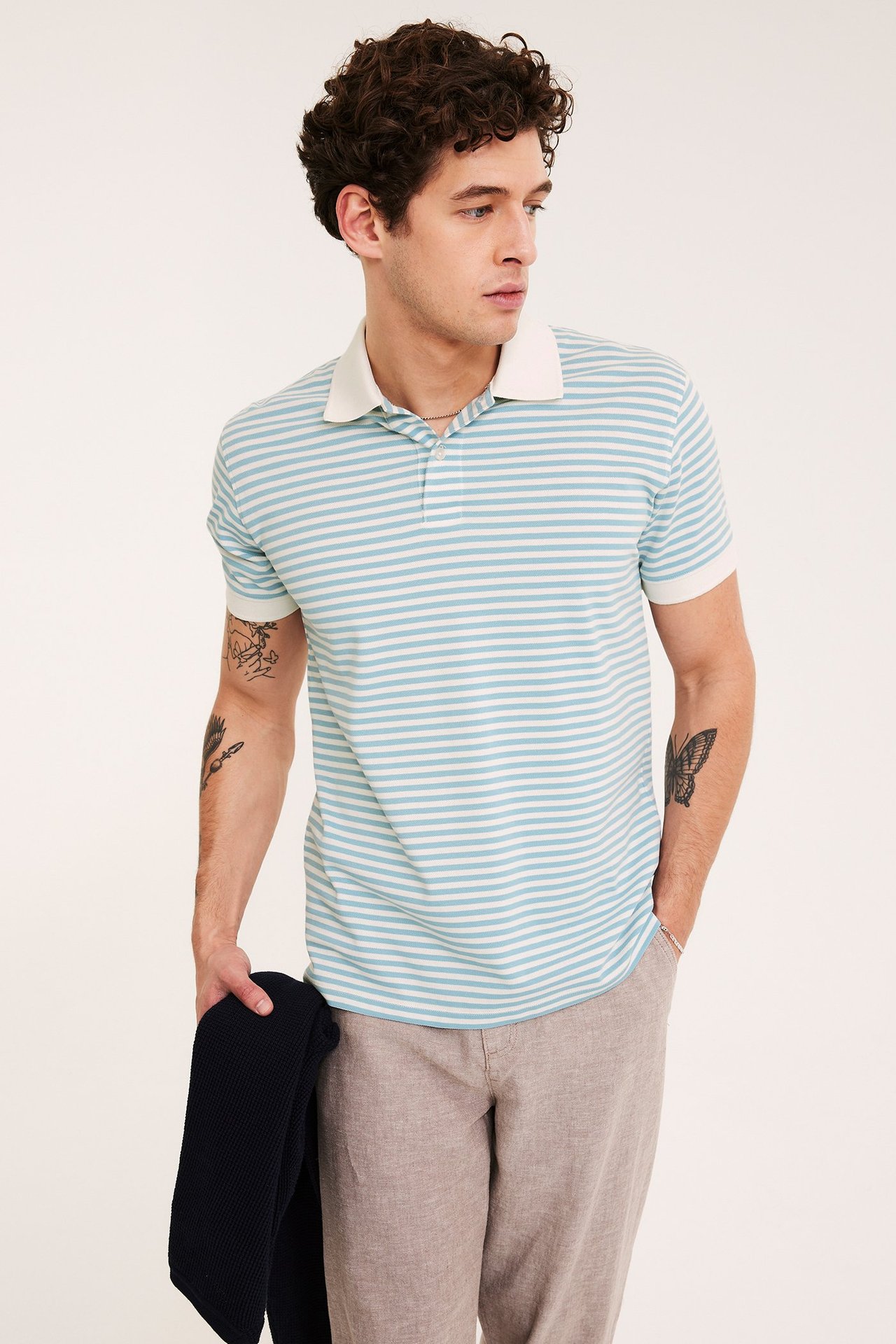 Framifrån: man i ljusblårandig bomullspiké, beigefärgade byxor, håller i en mörkblå stickad tröja.