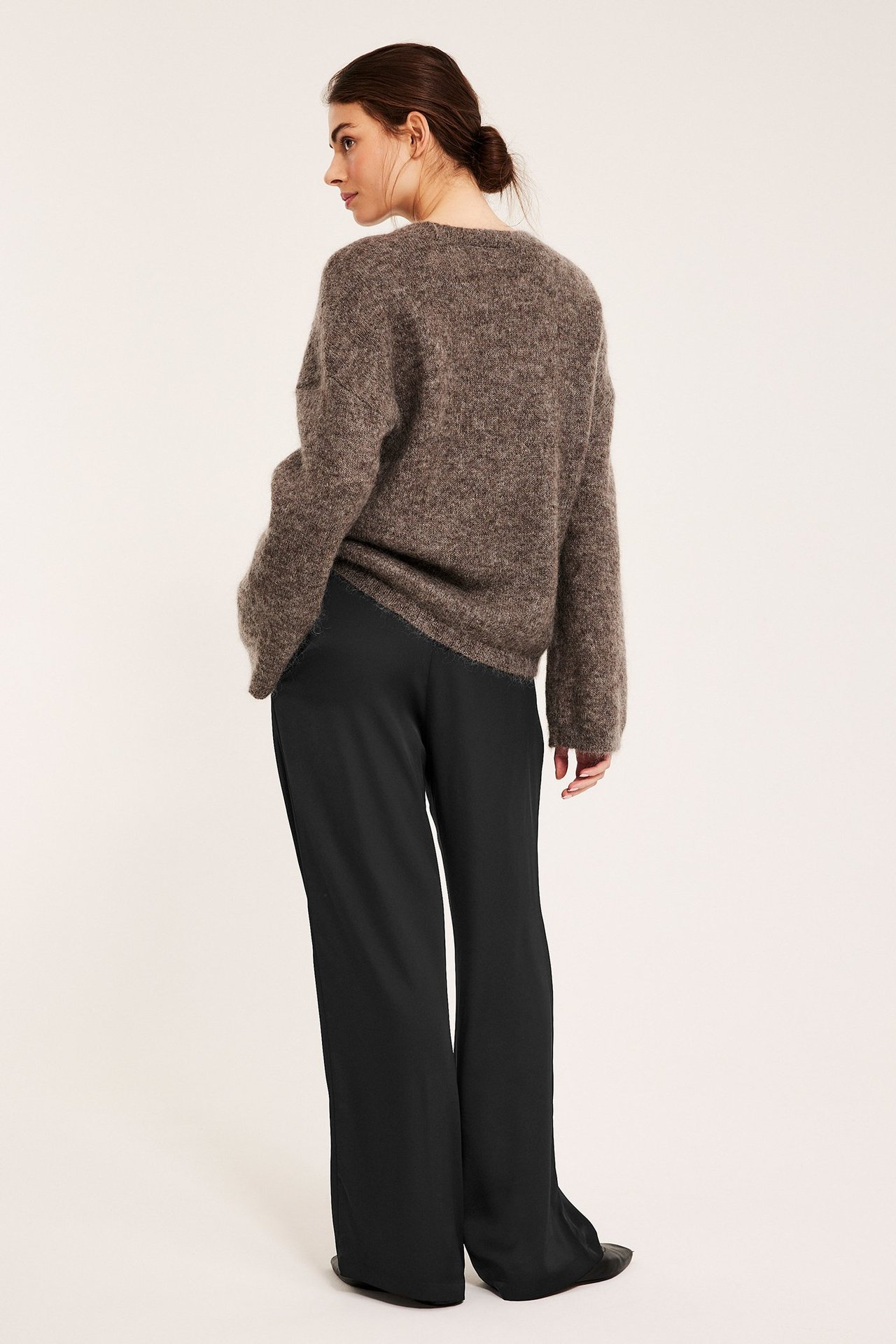 Rygg-sidevisning av kvinne i svarte vide satengbukser med strikk og en brun lodden genser.