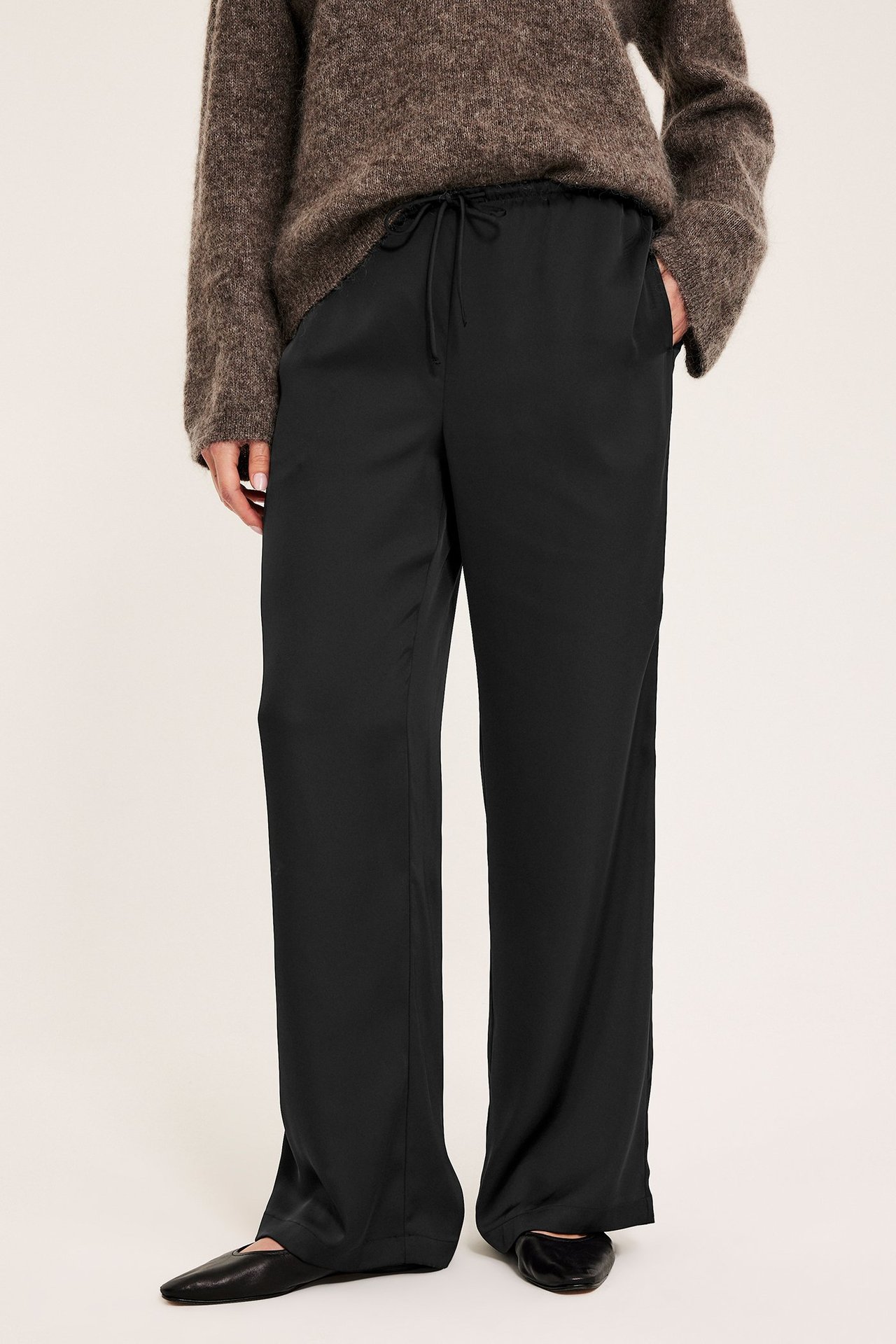 Forfra: kvinne i svarte vide satengbukser med snøring i midjen og en brun strikkegenser.