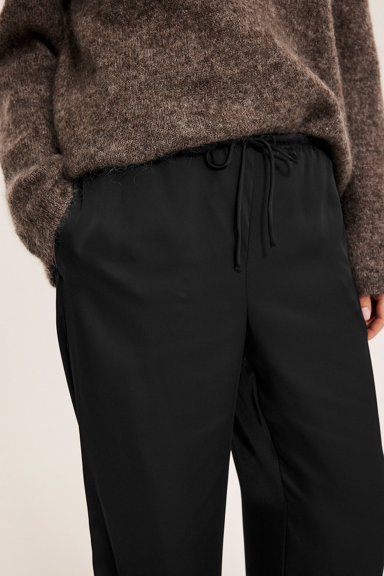 Nærbilde forfra: Kvinnens svarte satengbukser med snøring og sidelommer, vist med en brun genser.