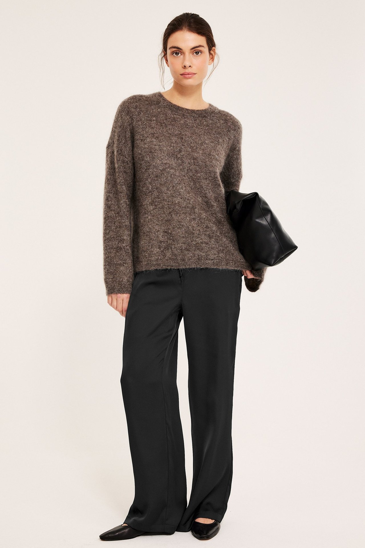 Frontbilde av kvinne i svarte vide satengbukser, brun strikkegenser og svarte ballerinasko, holder en veske.