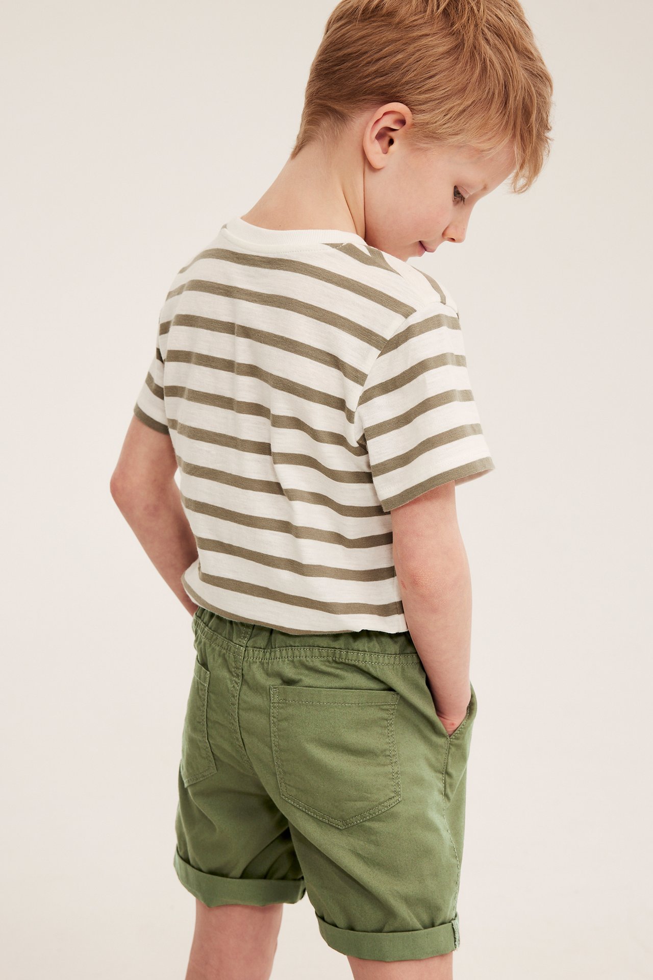 Chłopiec w kremowo-beżowym pasiastym T-shircie i zielonych bawełnianych szortach, z boku.