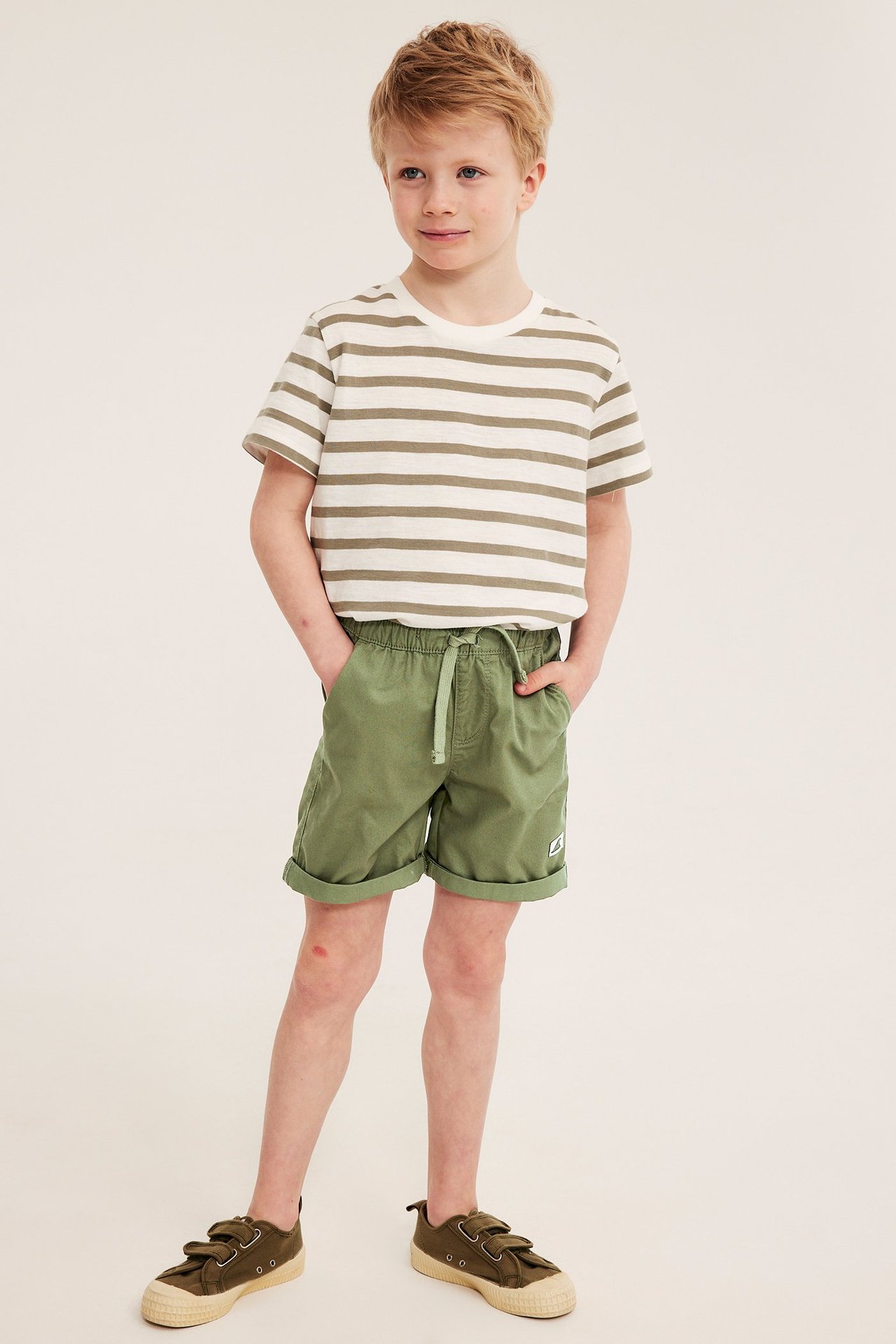 Edestäpäin: lapsi tummanvihreissä puuvillashortseissa, valkoraitaisessa paidassa ja tummanvihreissä kengissä.