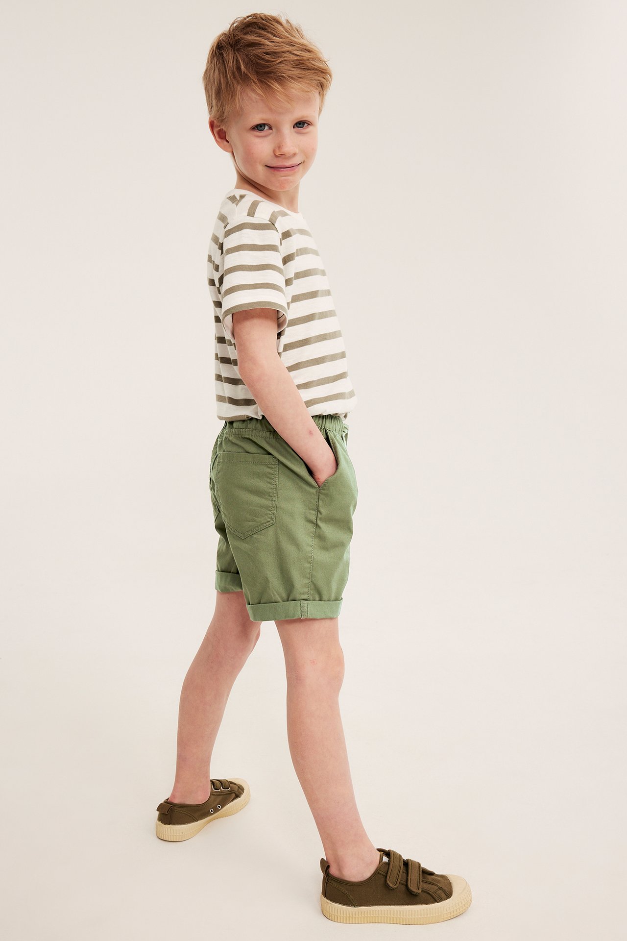 Chłopiec z boku w ciemnozielonych szortach bawełnianych, pasiastym t-shircie i oliwkowych sneakersach na rzepy.