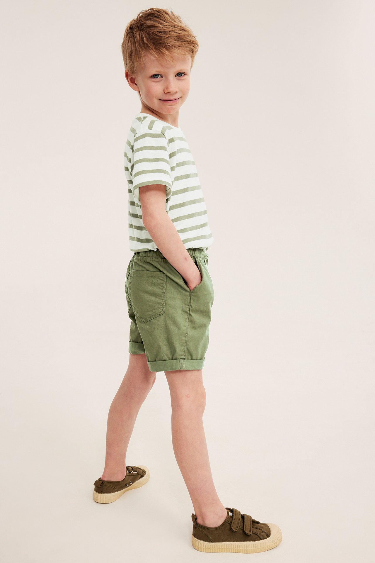 Widok z boku dziecka w ciemnozielonych bawełnianych szortach, koszulce w paski i zielonych trampkach.
