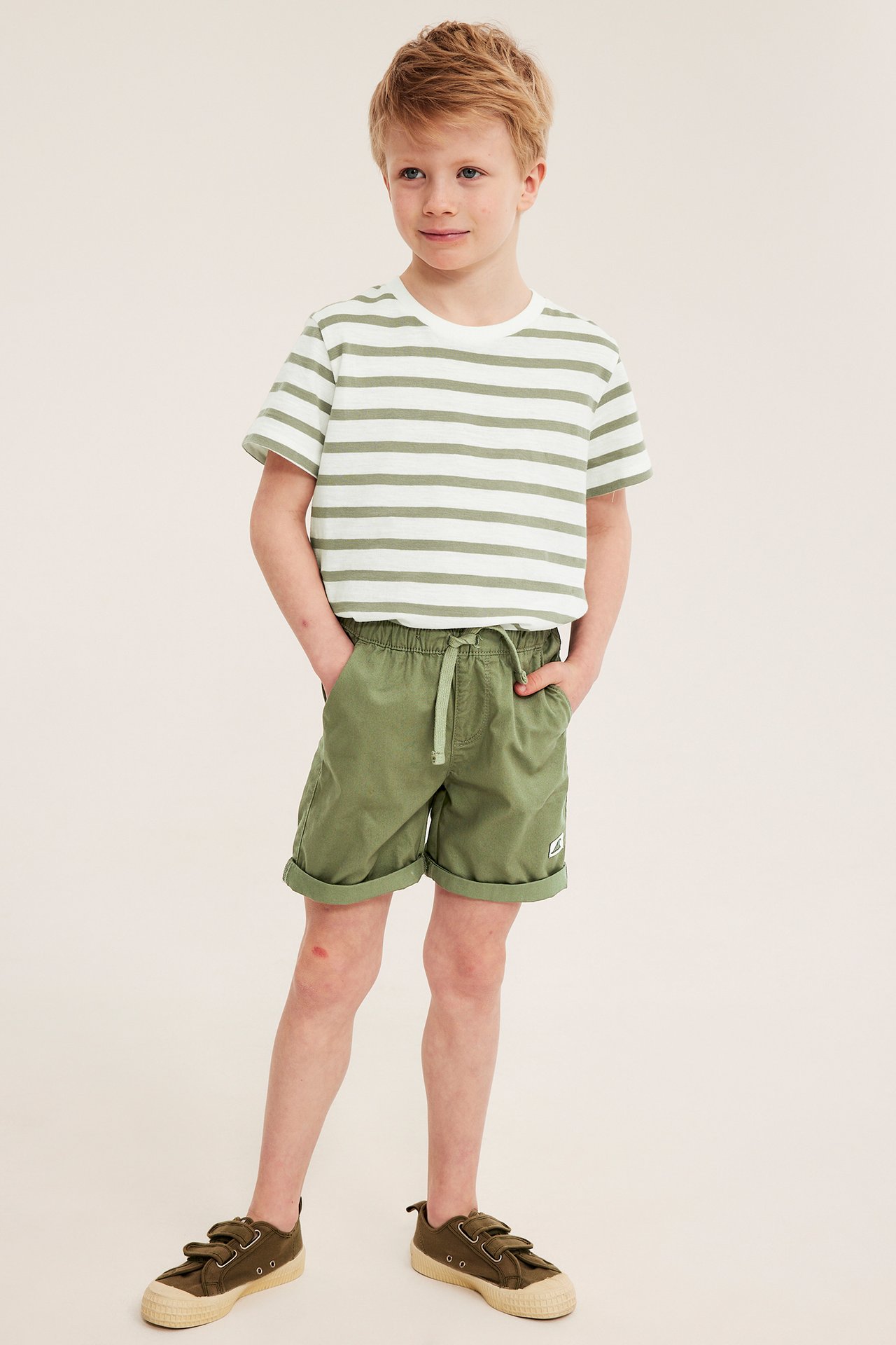 Front view av ett barn i mörkgröna bomullstwillshorts med dragsko, randig t-shirt och mörkgröna skor.
