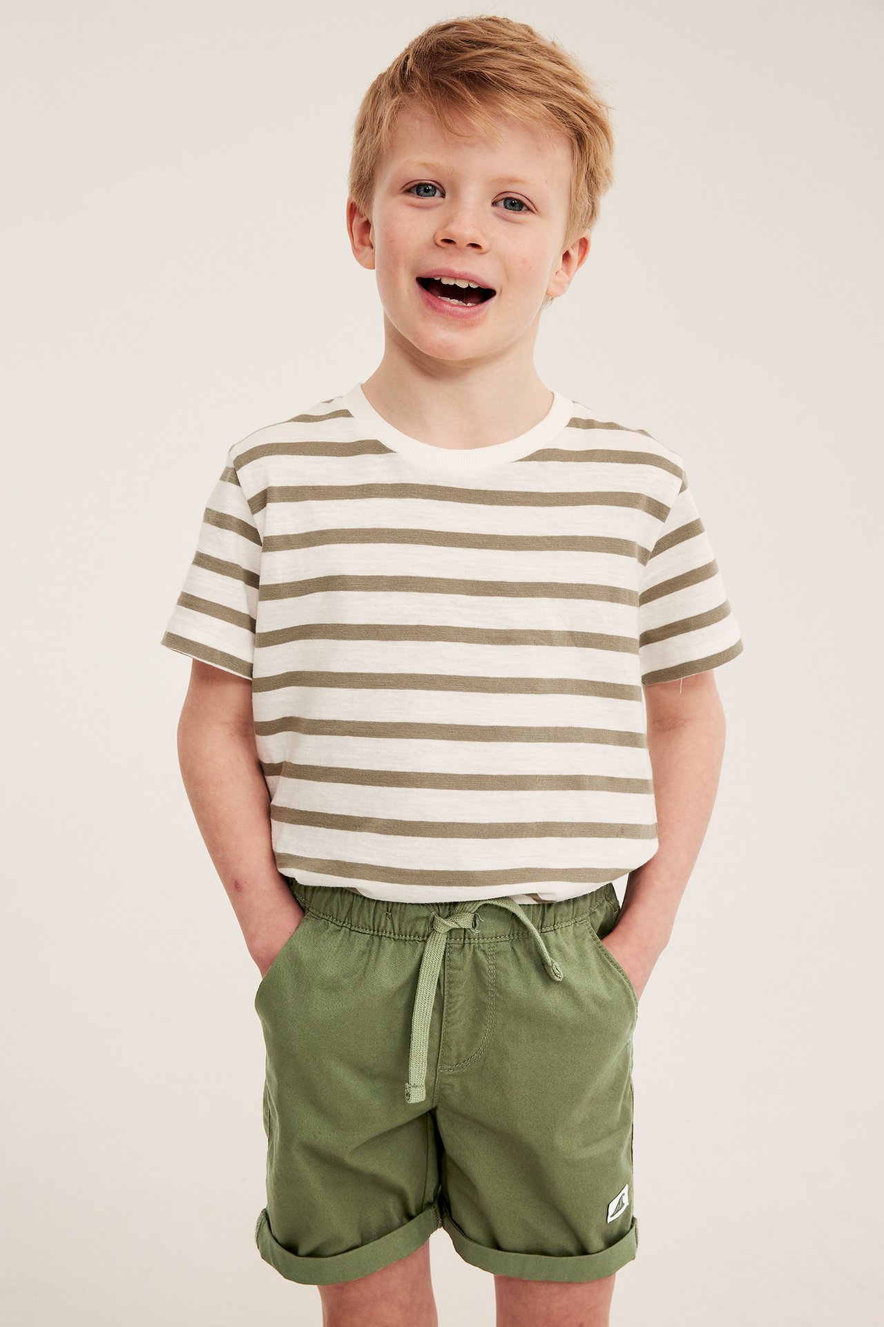 Etunäkymä: Lapsella on yllään tummanvihreät puuvillatwillishortsit kiristysnyörillä ja raidallinen t-paita.