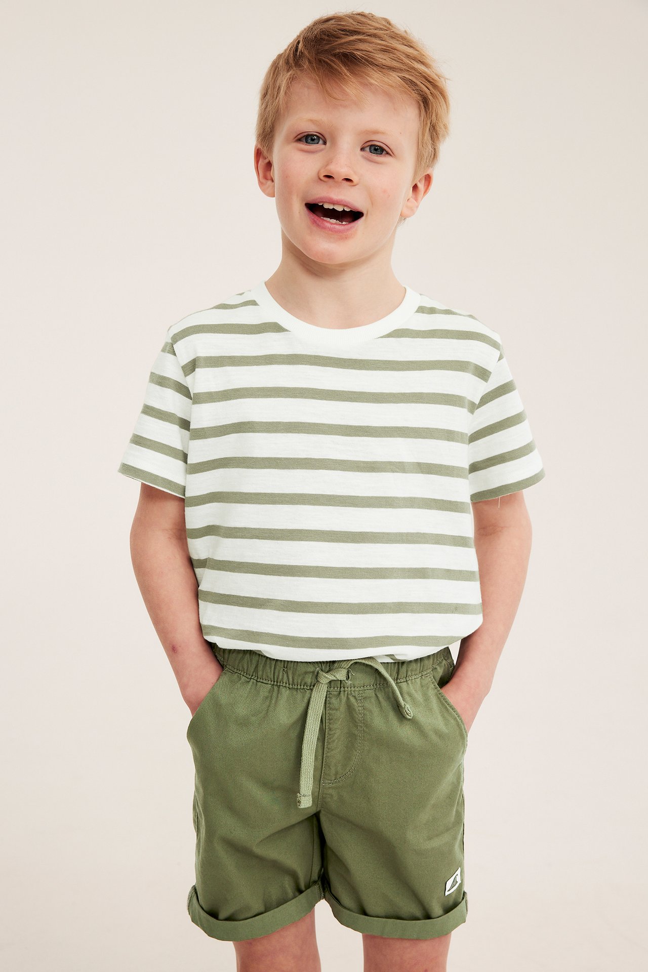 Etunäkymä: Lapsi tummanvihreissä puuvillatwillishortseissa, joustovyötäröllä ja raidallisessa t-paidassa.