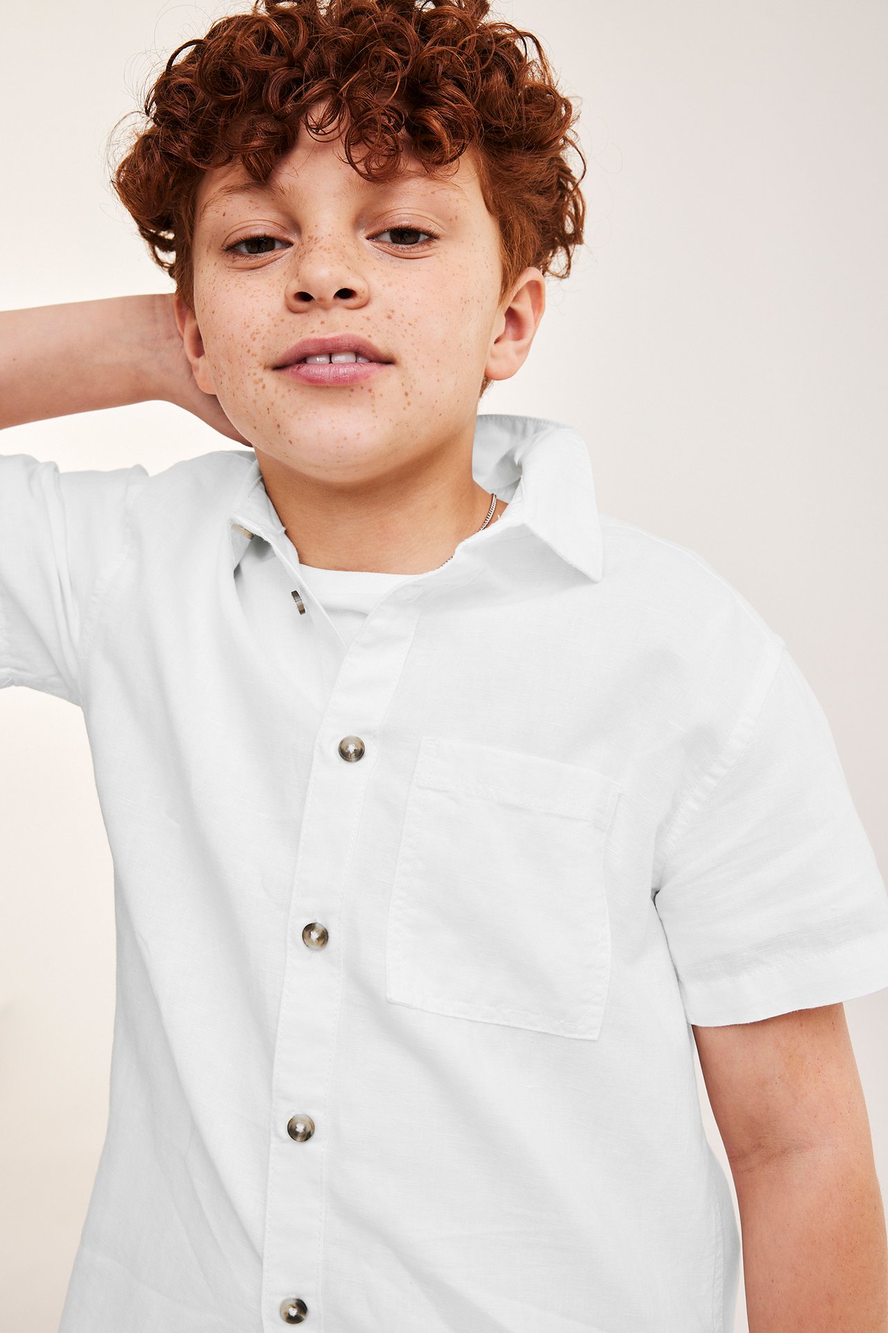Lähikuva edestä lapsesta, yllään valkoinen pellavasekoitteinen lyhythihainen paita ja vaaleat napit, t-paidan päällä.