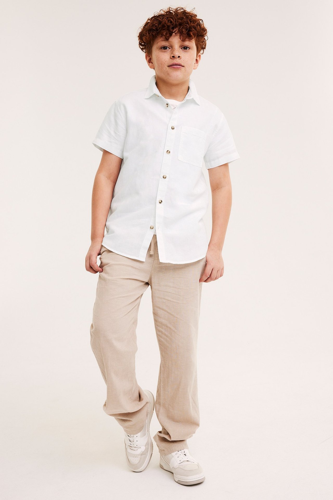 Widok z przodu dziecka w białej lnianej koszuli, beżowych spodniach ze sznurkiem i jasnych sneakersach.