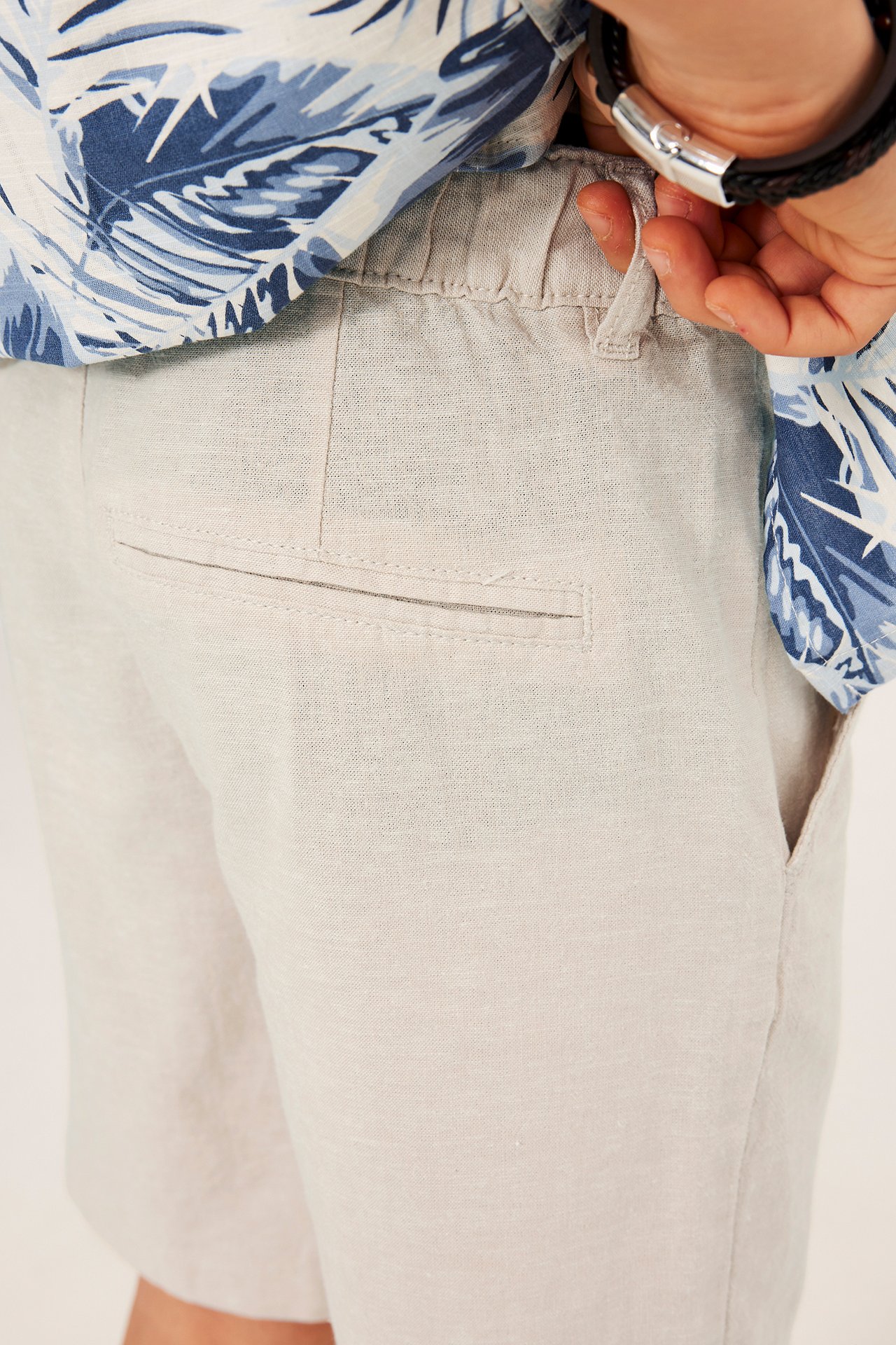 Nærbilde bakfra av barn i lysebeige linshorts med strikk i midjen, mønstret skjorte og et armbånd.