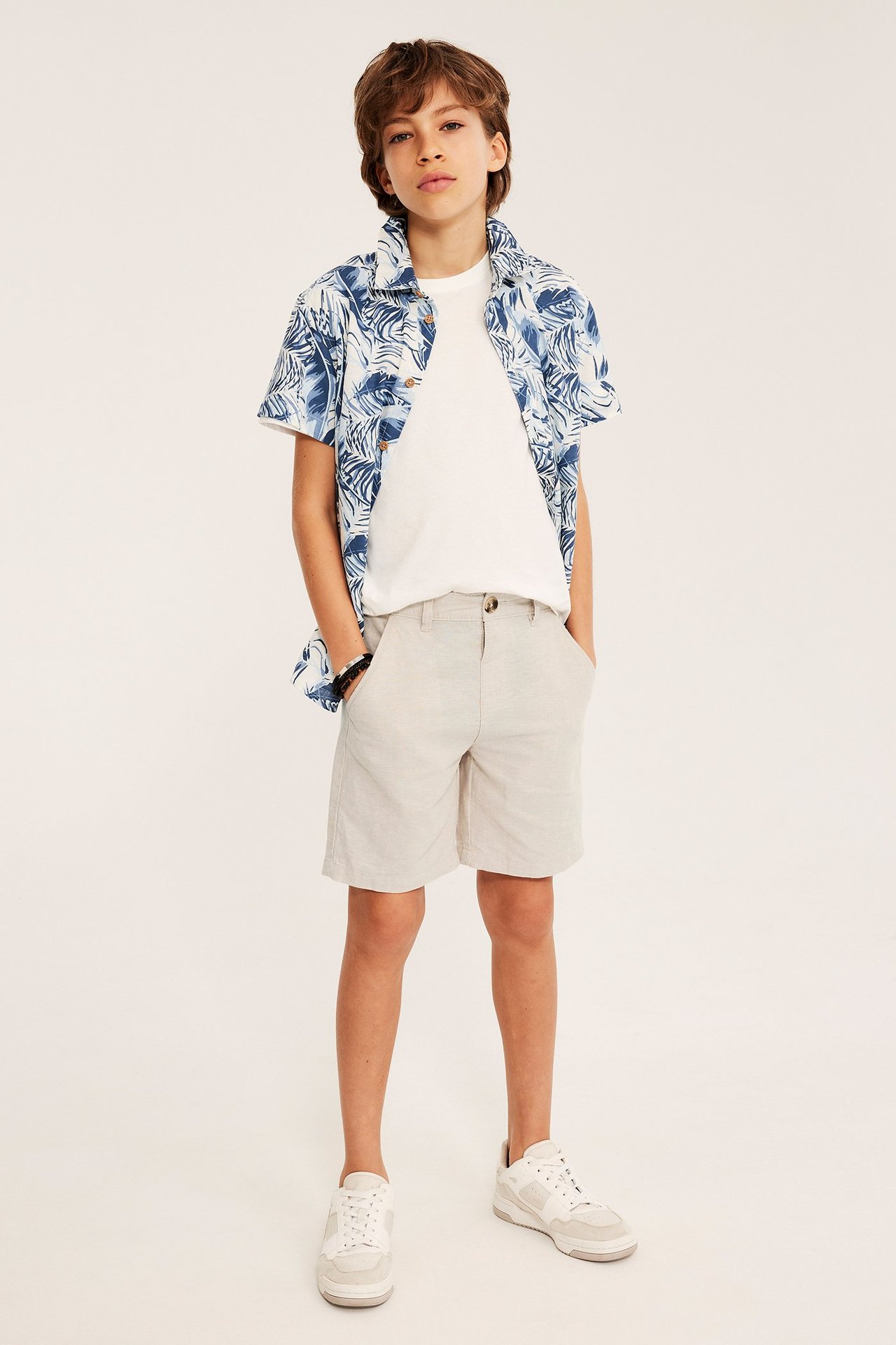 Edestä: lapsi vaaleabeigeissä pellavasekoiteshortseissa, valkoisessa t-paidassa ja avoimessa sinikuviollisessa paidassa.