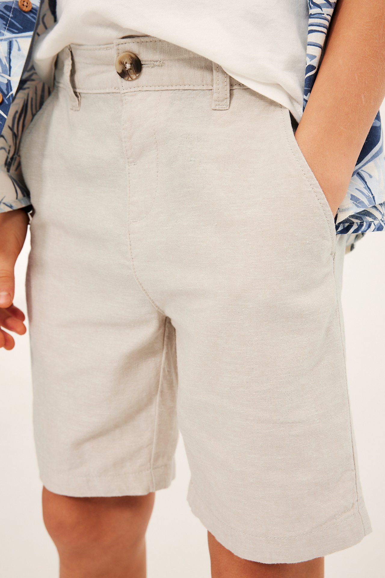 Frontbild av barn i ljusbeige linneshorts, knappstängning, med vit t-shirt och blå mönstrad skjorta.